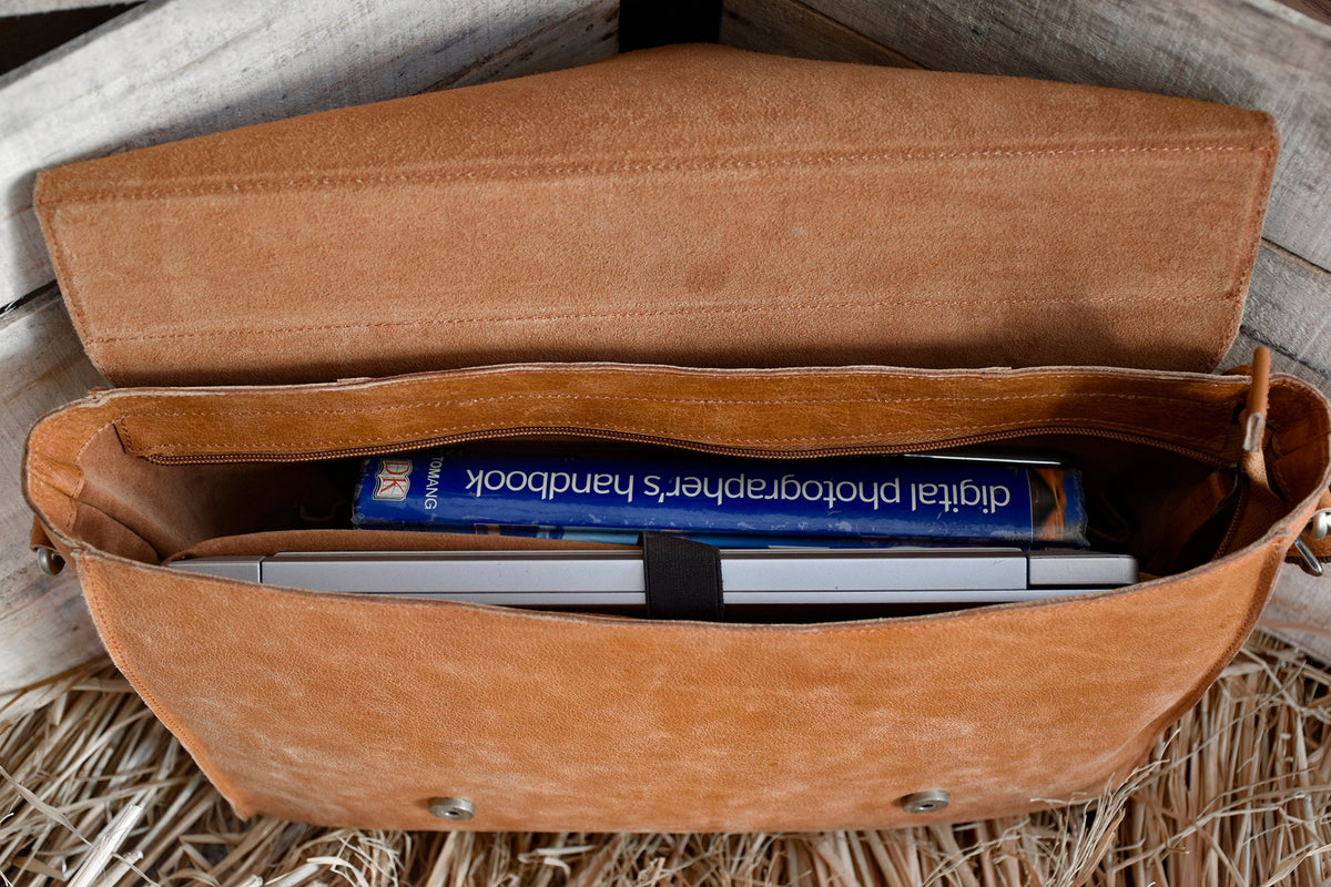 May include: A brown leather messenger bag with a zippered pocket and a flap closure. The bag is open and a laptop and a book titled "Digital Photographer's Handbook" are inside.