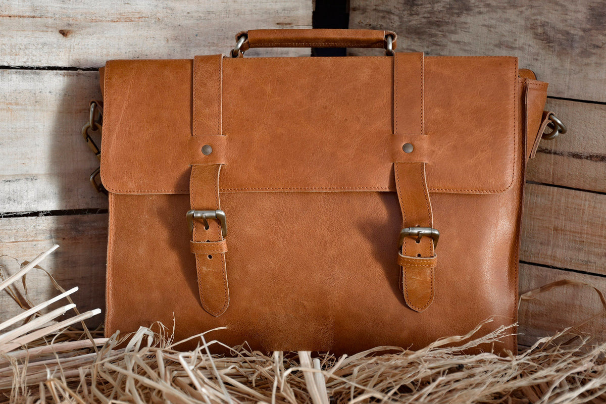 May include: A brown leather briefcase with two buckles and a handle. The briefcase is sitting on a bed of straw.
