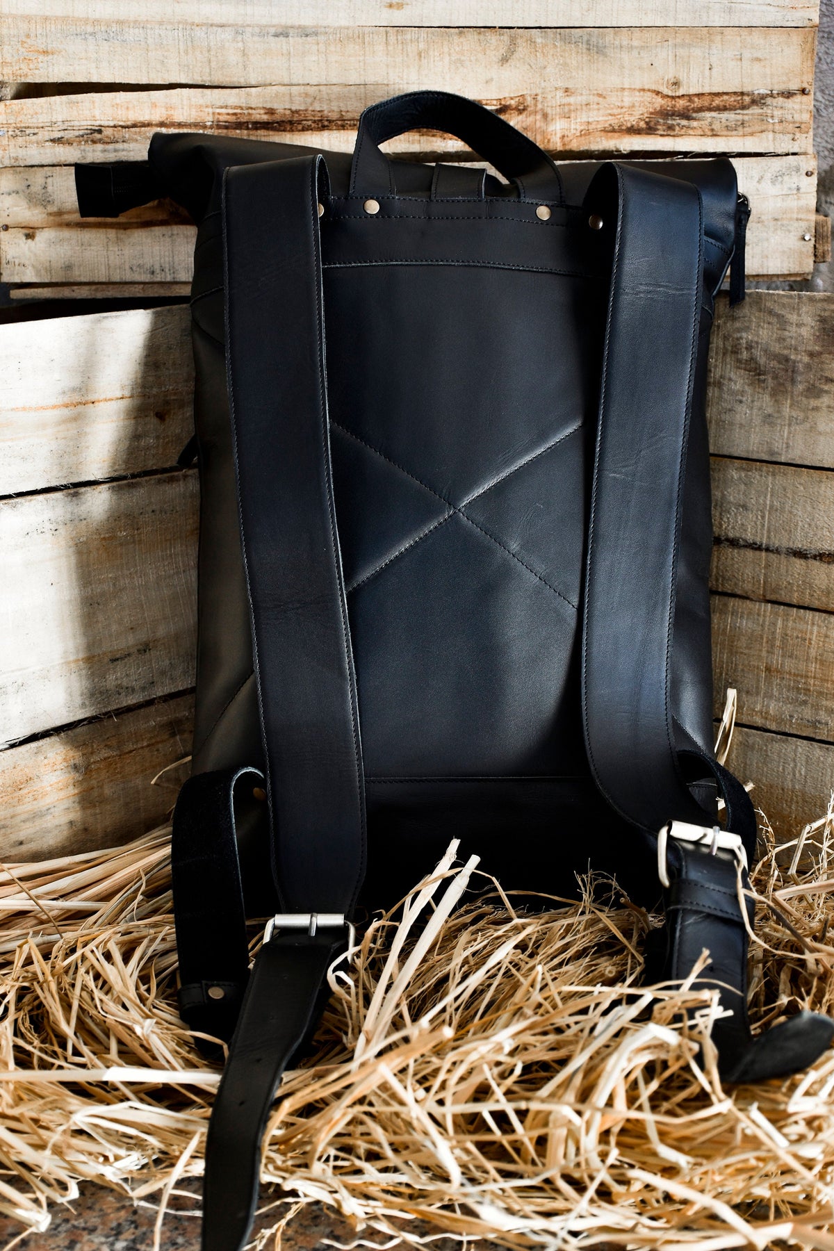 May include: A black leather backpack with adjustable straps and a roll-top closure. The backpack has a geometric design on the back panel and is sitting on a bed of straw.