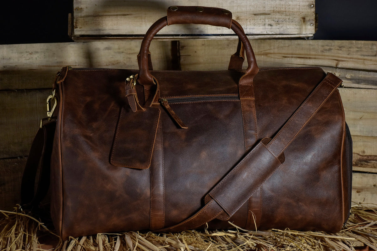 May include: A brown leather duffel bag with a top handle and a shoulder strap. The bag has a zippered closure and a small pocket on the front.