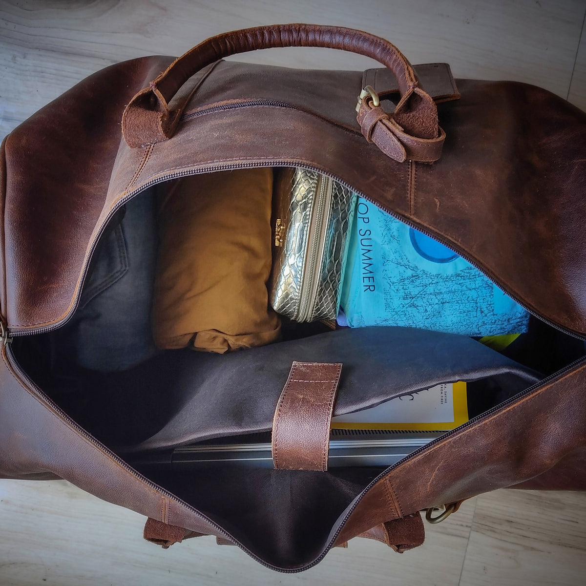 May include: A brown leather duffel bag with a zipper closure. The bag is open and filled with various items, including a blue and white patterned fabric, a brown fabric pouch, a silver and black patterned pouch, and a black laptop case.