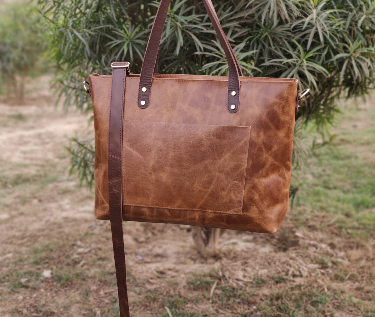 May include: A brown leather tote bag with a brown leather strap. The bag has a front pocket and a top zipper closure.