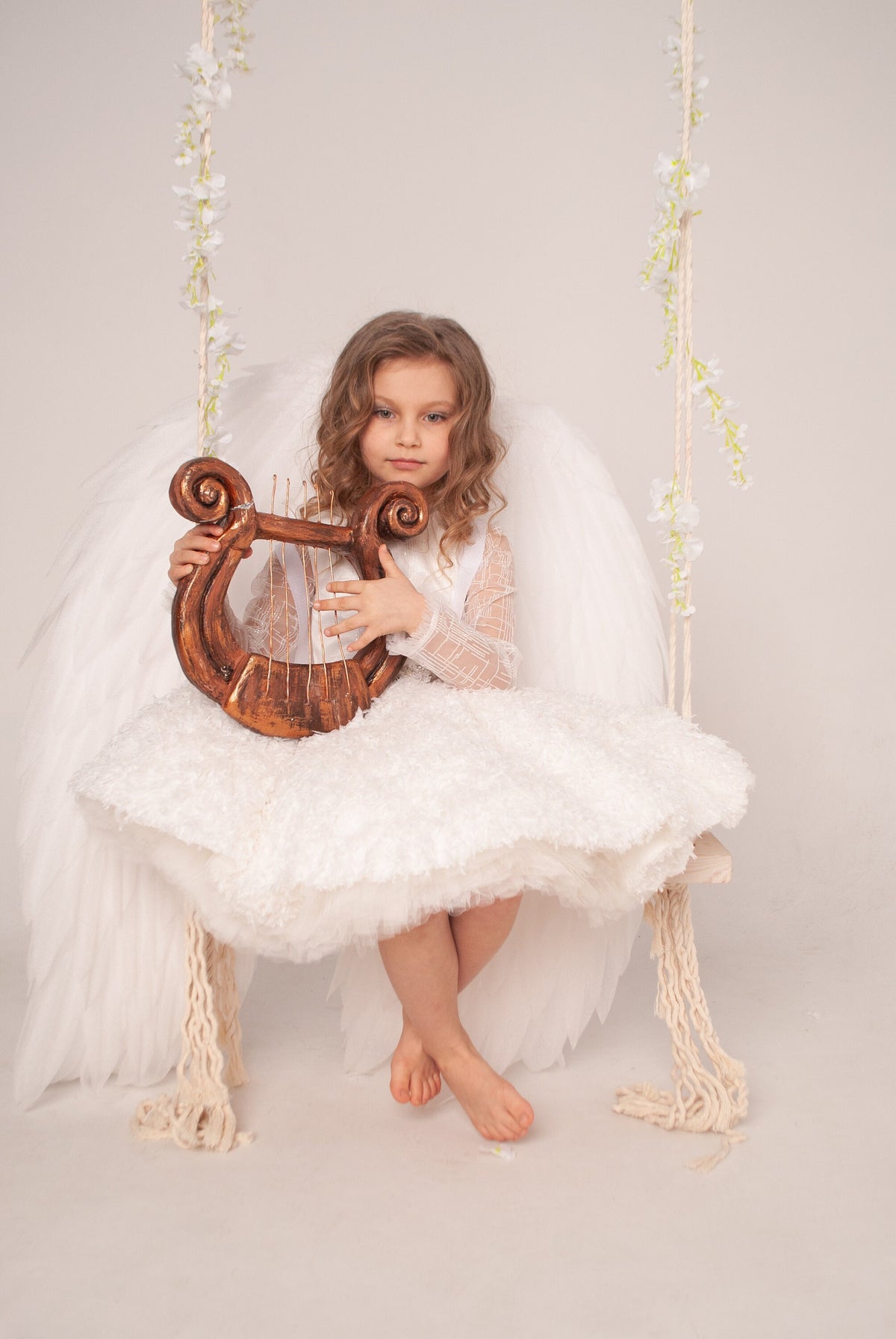 May include: A child with wavy hair wearing a white textured dress and large white feathered wings, sitting on a wooden swing decorated with white floral vines, holding a small brown wooden lyre with thin vertical strings in their hands.