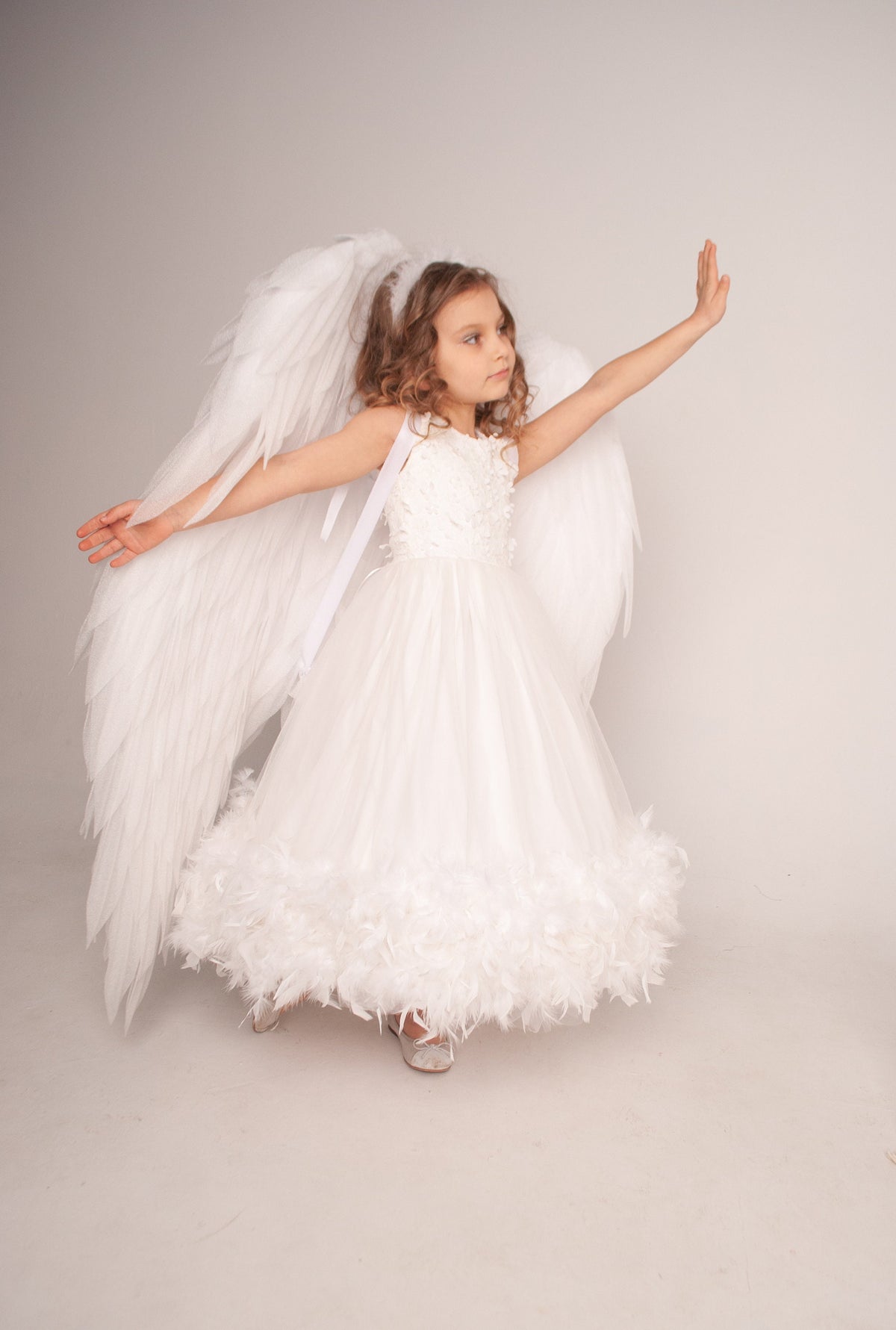 May include: A white floor-length dress featuring a bodice with floral embellishments, a layered tulle skirt with a feathered hemline, and large white feathered angel wings attached to the back with white ribbon straps, worn with silver shoes.