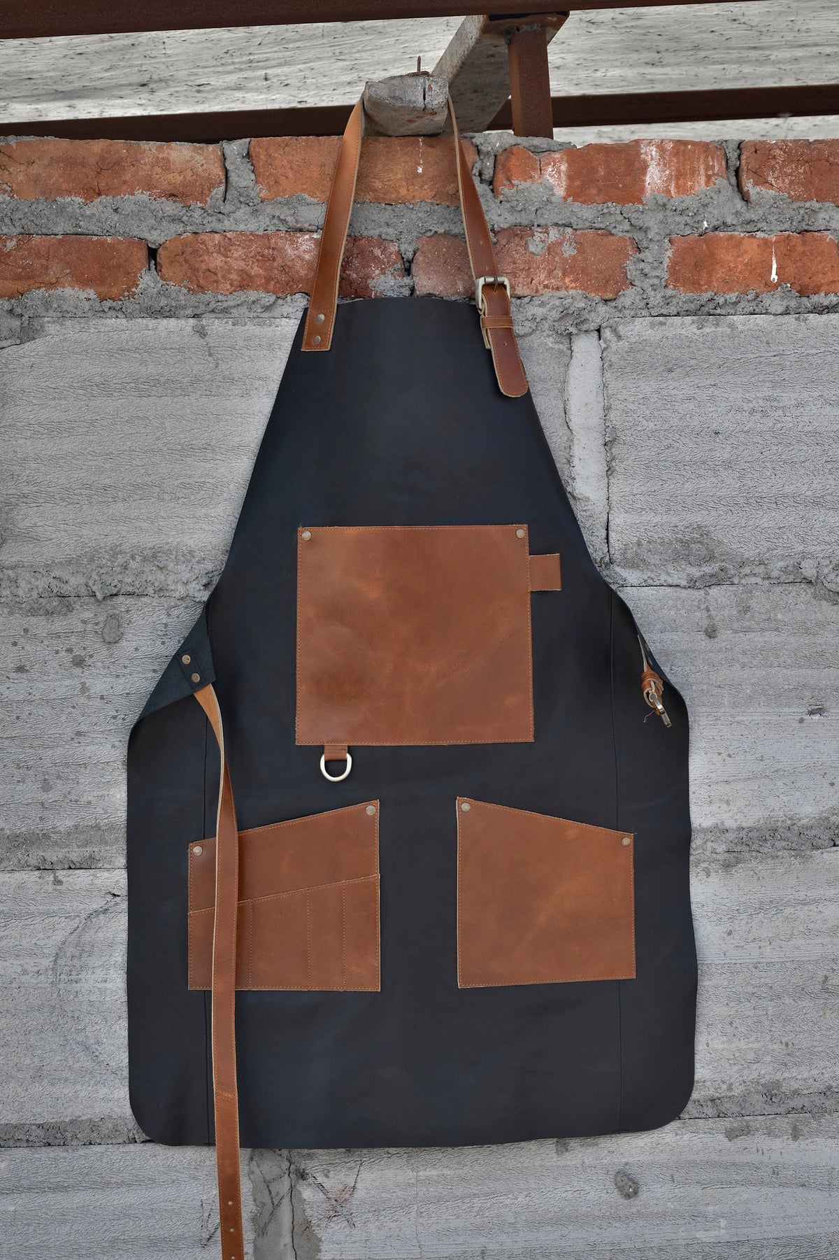 May include: A black canvas apron with brown leather straps, pockets, and trim. The apron has a large pocket on the front and two smaller pockets on the sides.