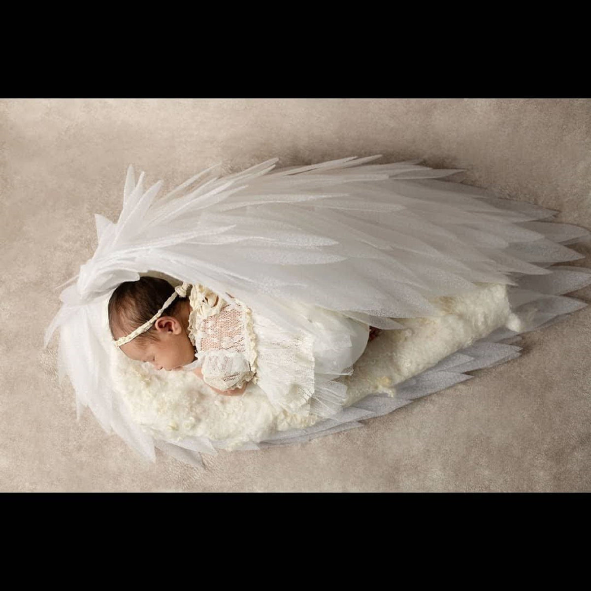 May include: A newborn baby wearing a white lace dress and a white headband, sleeping peacefully under a pair of large white angel wings.