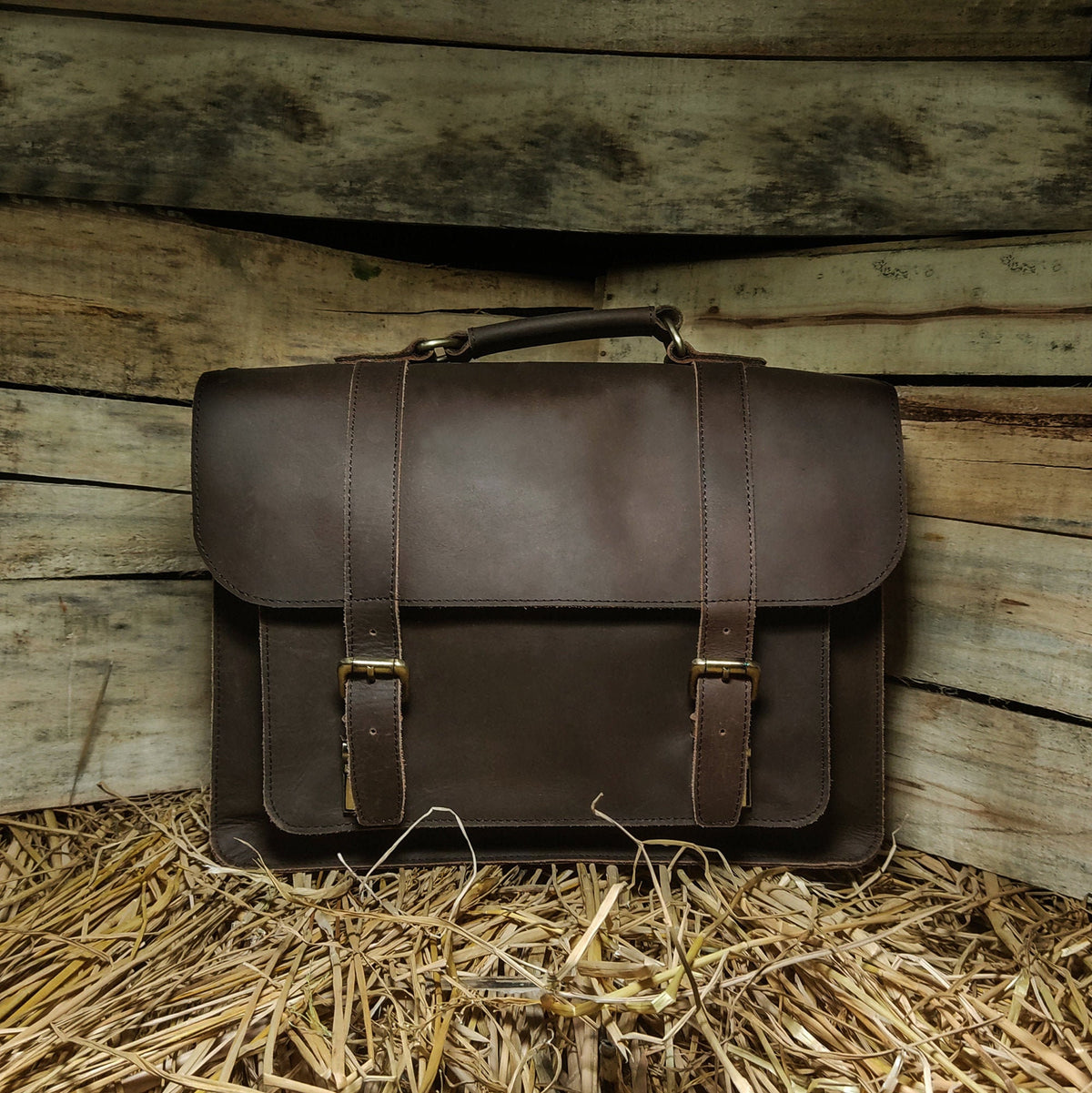 May include: A brown leather briefcase with two buckles and a handle. The briefcase is sitting on a bed of straw.