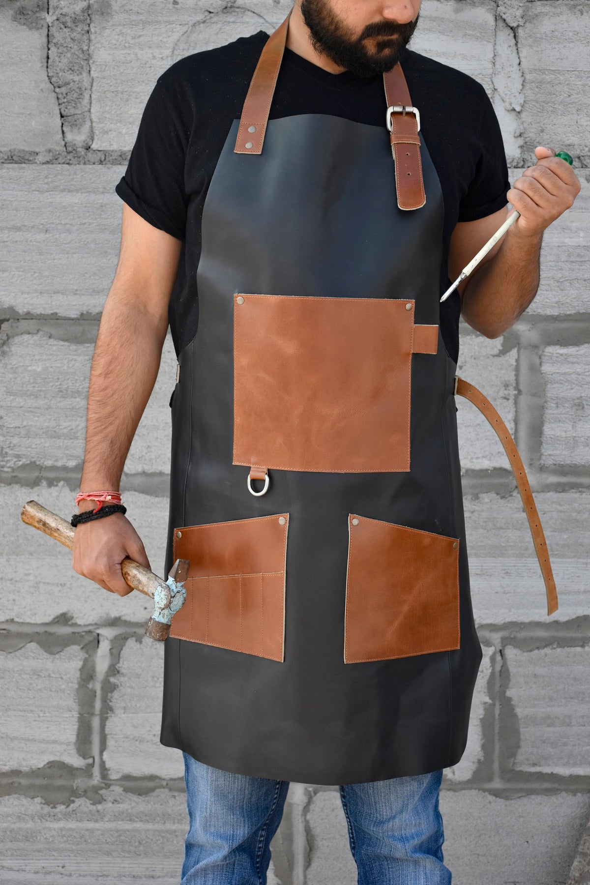 May include: A black leather apron with brown leather accents and multiple pockets. The apron has a large pocket on the front with a metal ring. The apron is being worn by a person holding a hammer.