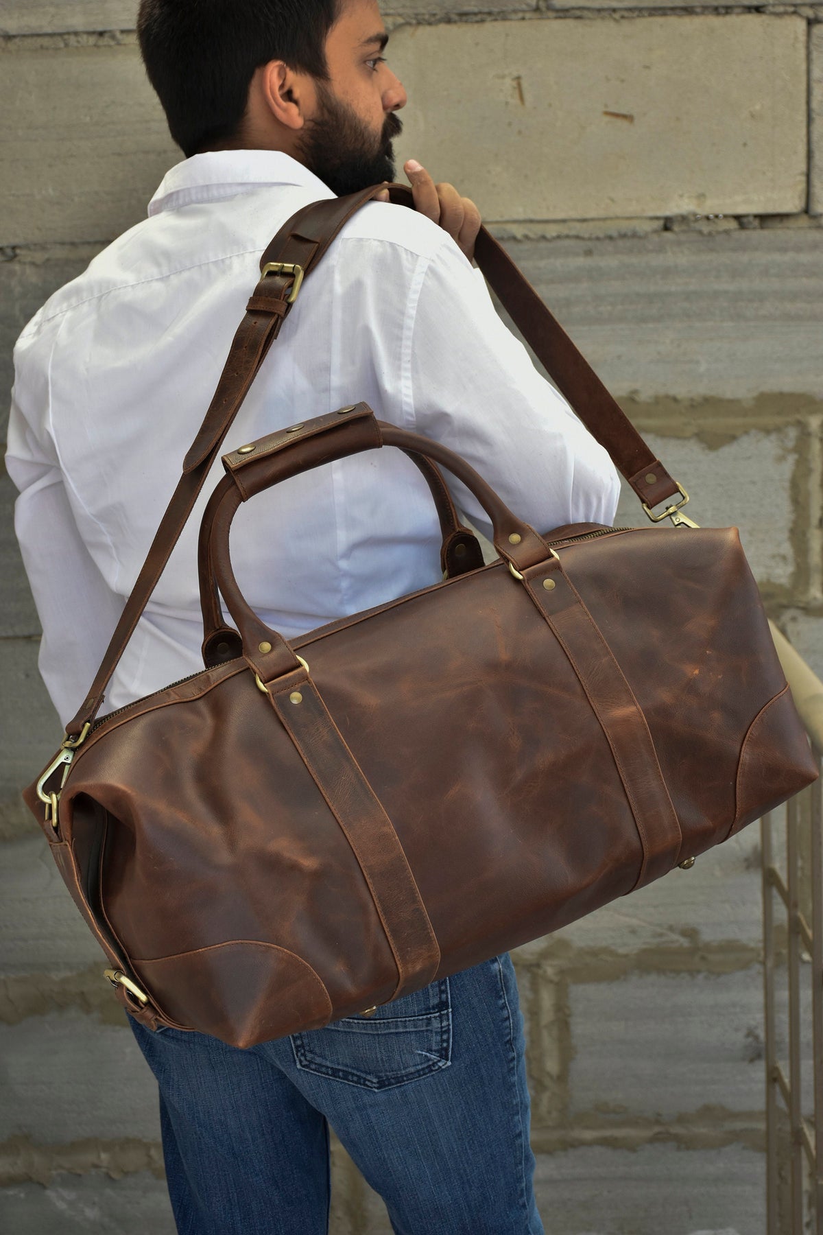 May include: A brown leather duffel bag with a long strap and brass hardware. The bag is being carried by a person wearing a white shirt and blue jeans.