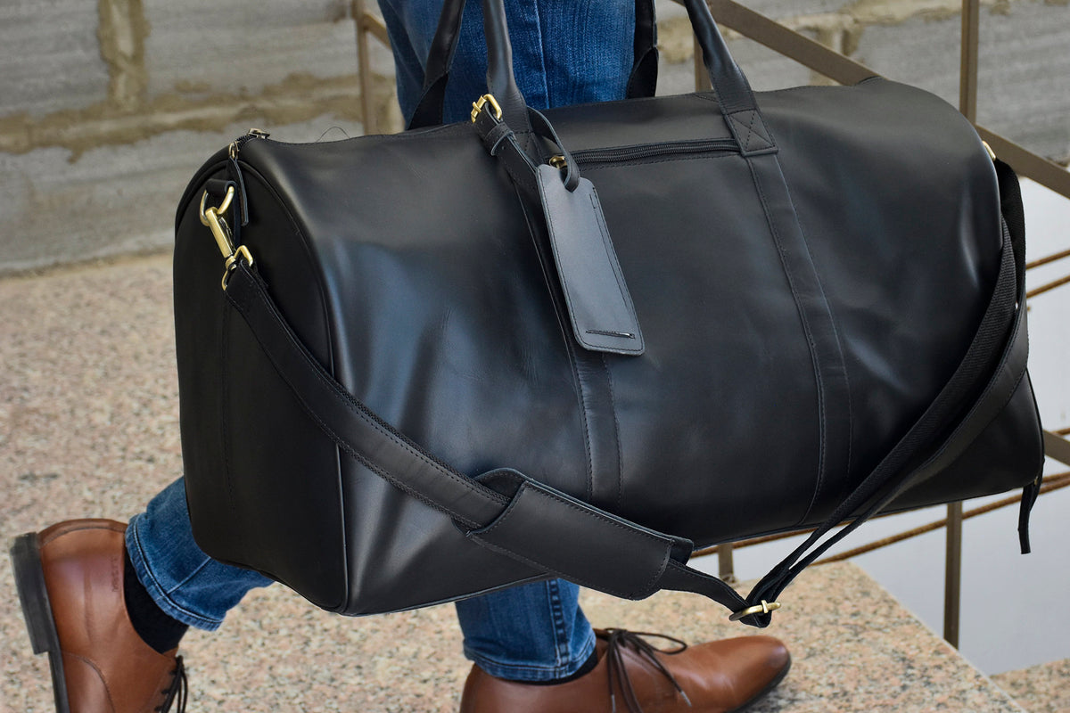May include: A black leather duffel bag with a gold zipper and a gold luggage tag. The bag has a long strap that can be worn over the shoulder.