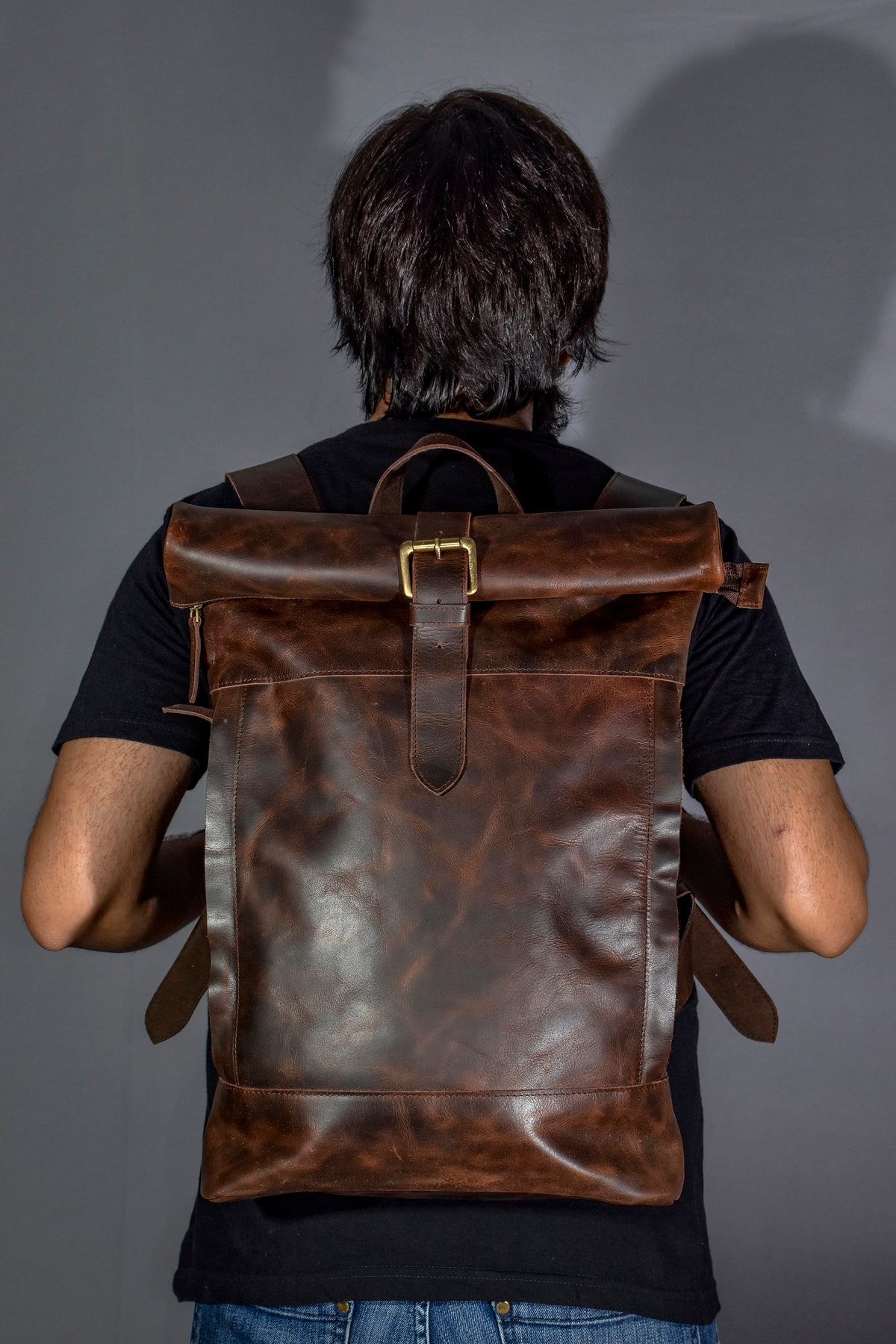 May include: A brown leather backpack with a roll-top closure and a buckle. The backpack has a distressed look and is worn by a person.