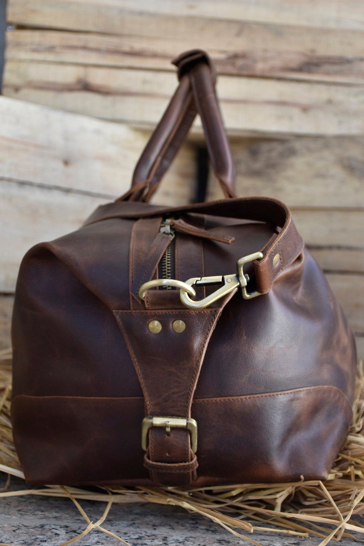 May include: A brown leather duffel bag with a brass buckle and a brass hook closure. The bag has a distressed look and is made of high-quality leather.