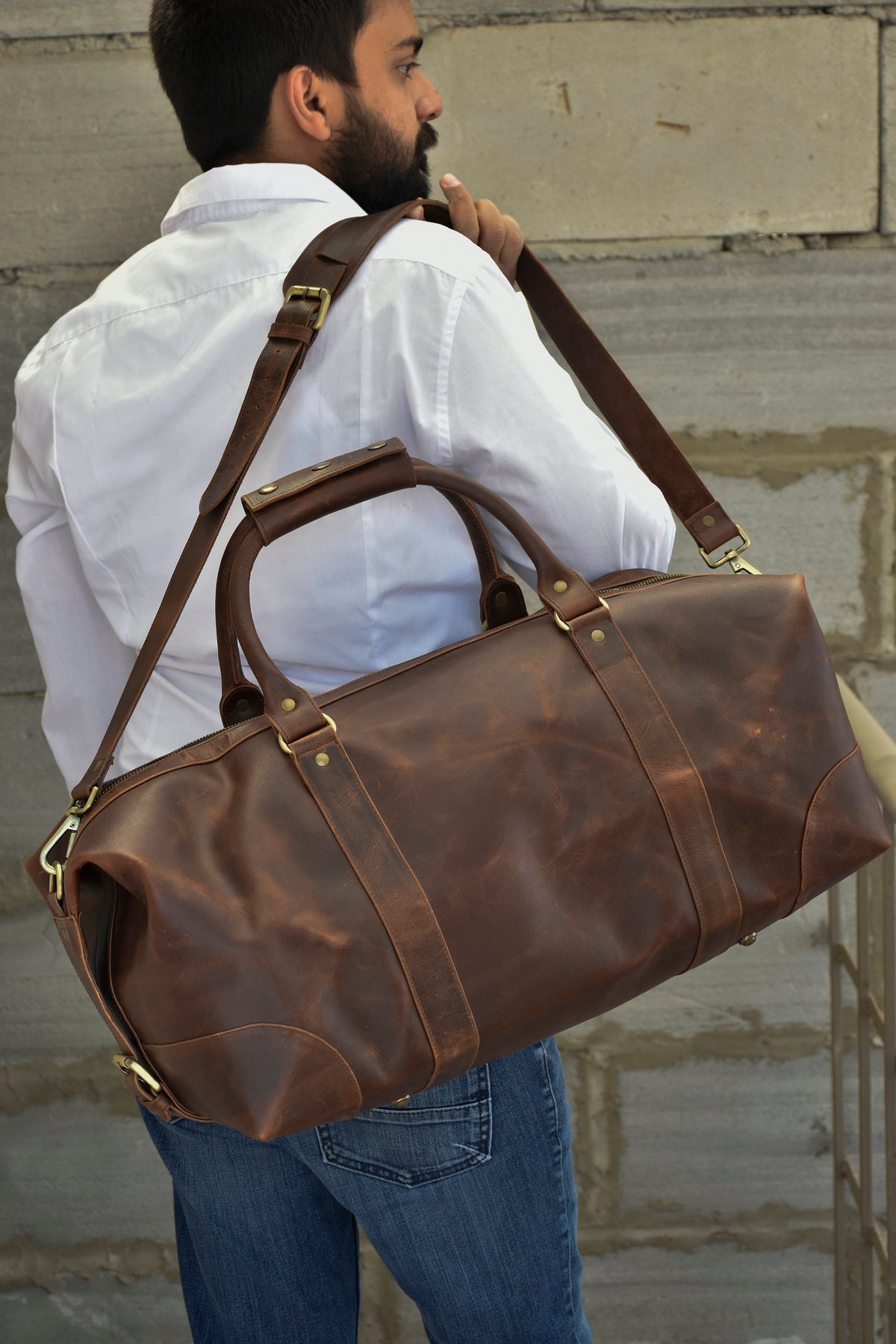 May include: A brown leather duffel bag with a long strap and brass hardware. The bag is being carried by a person wearing a white shirt and blue jeans.
