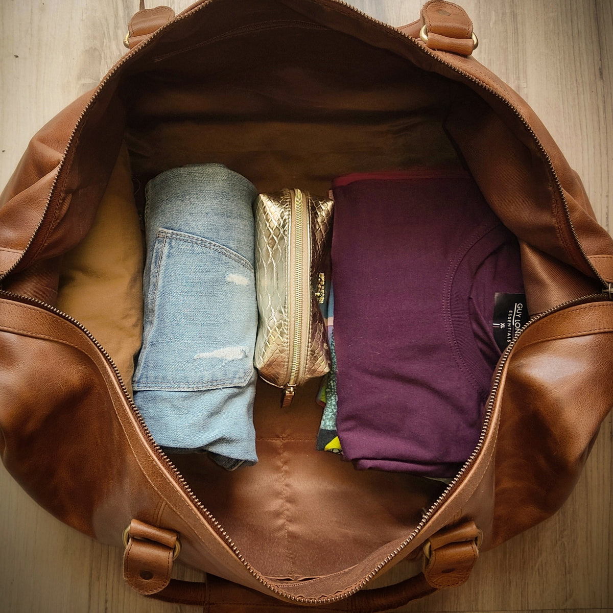 May include: A brown leather duffel bag with a zipper closure. The bag is open and filled with items, including a pair of blue jeans, a purple t-shirt, and a gold makeup bag.
