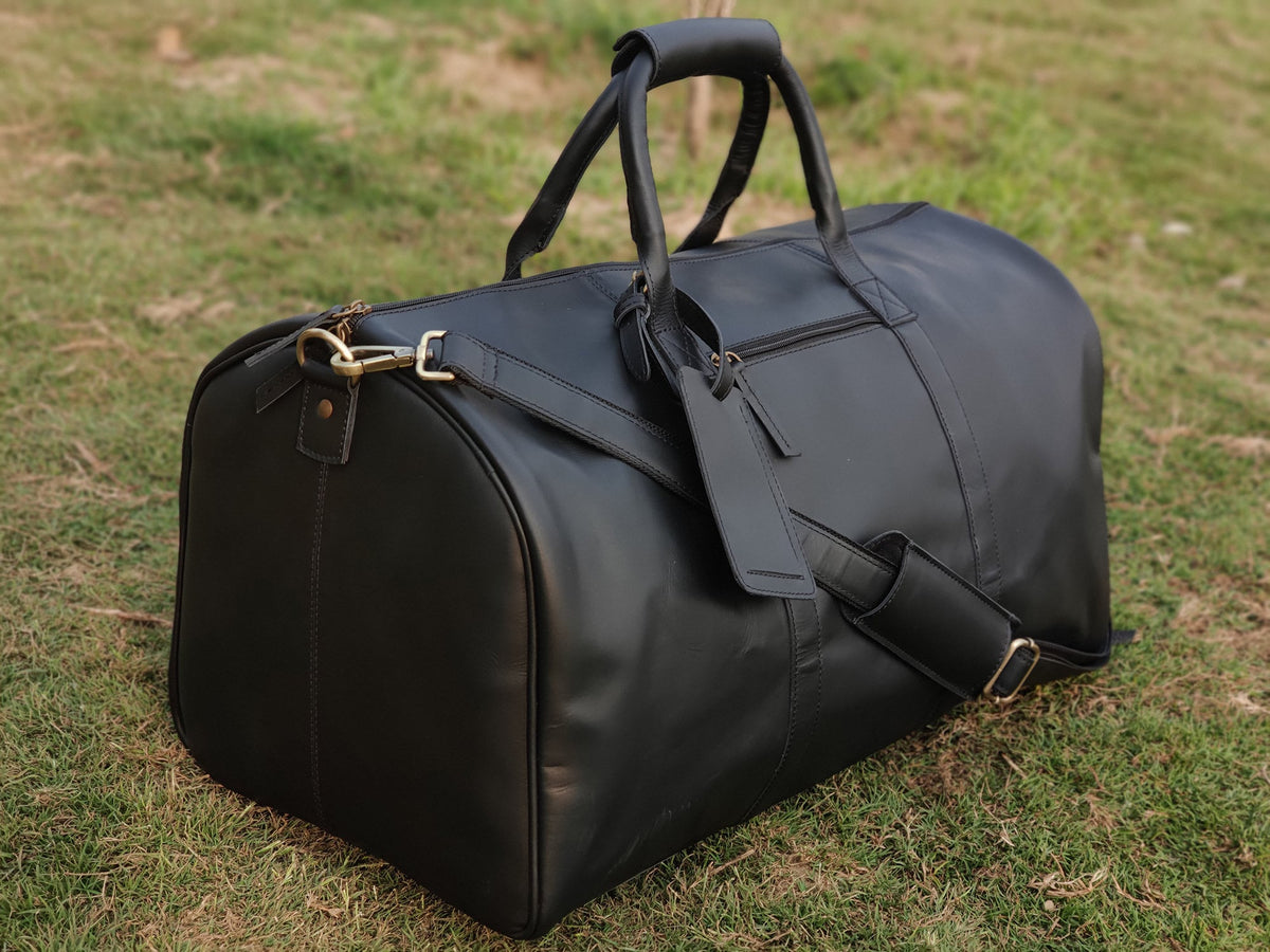 May include: A black leather duffel bag with two top handles and a shoulder strap. The bag has a zipper closure and a brass buckle.