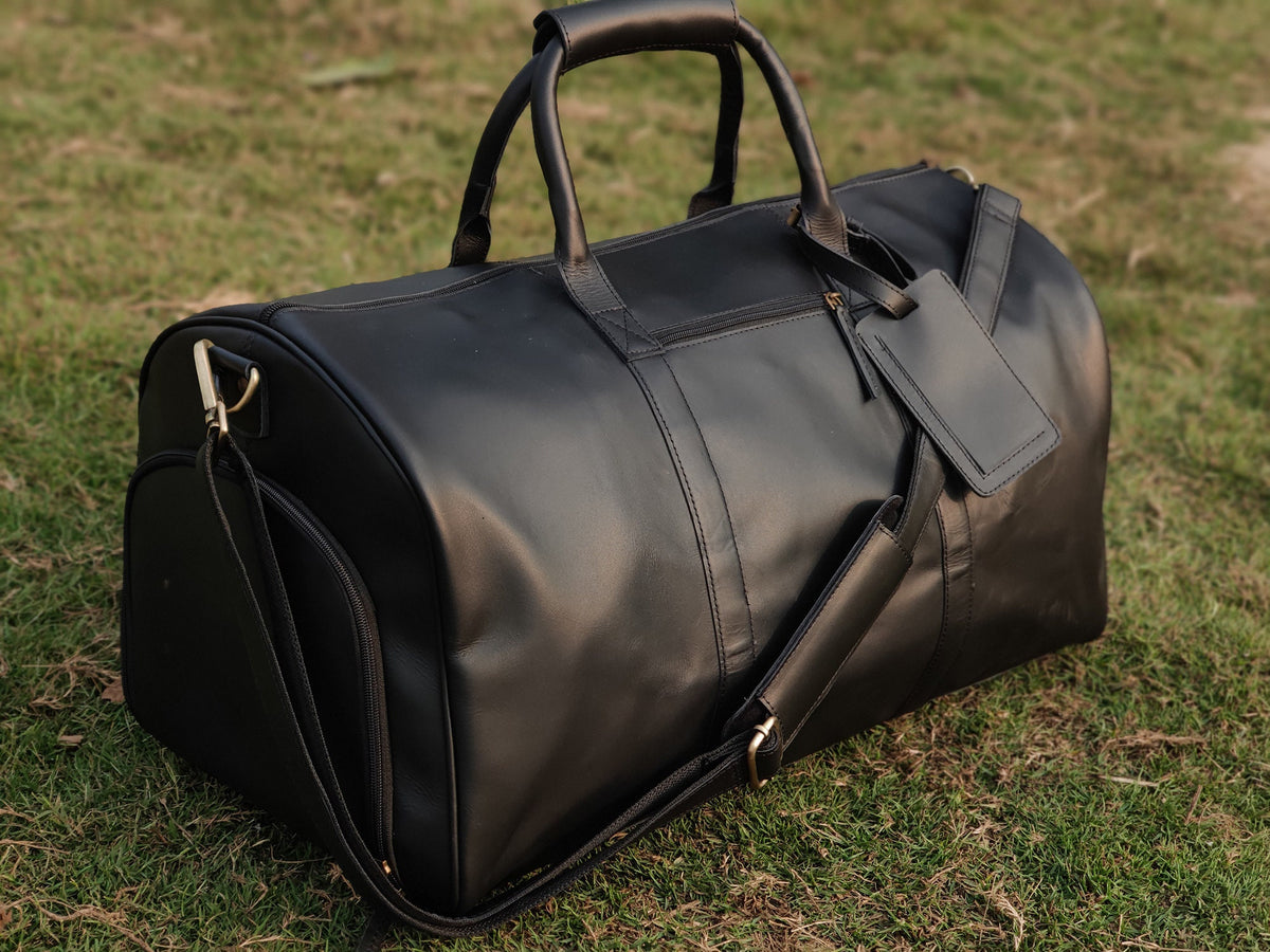 May include: A black leather duffel bag with a top handle and a shoulder strap. The bag has a zippered closure and a small leather tag attached to the zipper.