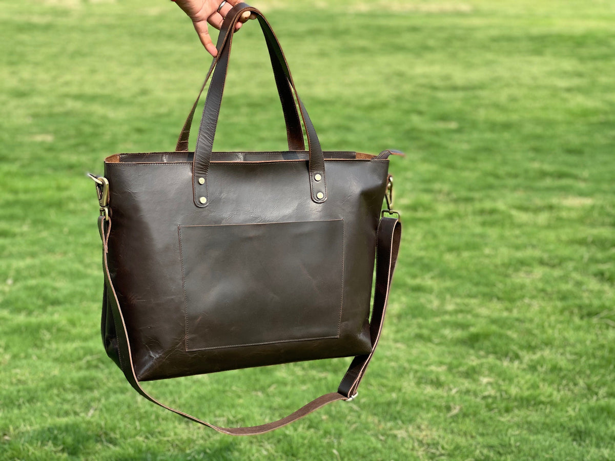May include: A brown leather tote bag with a long shoulder strap and a front pocket. The bag has brass hardware and a top zipper closure.