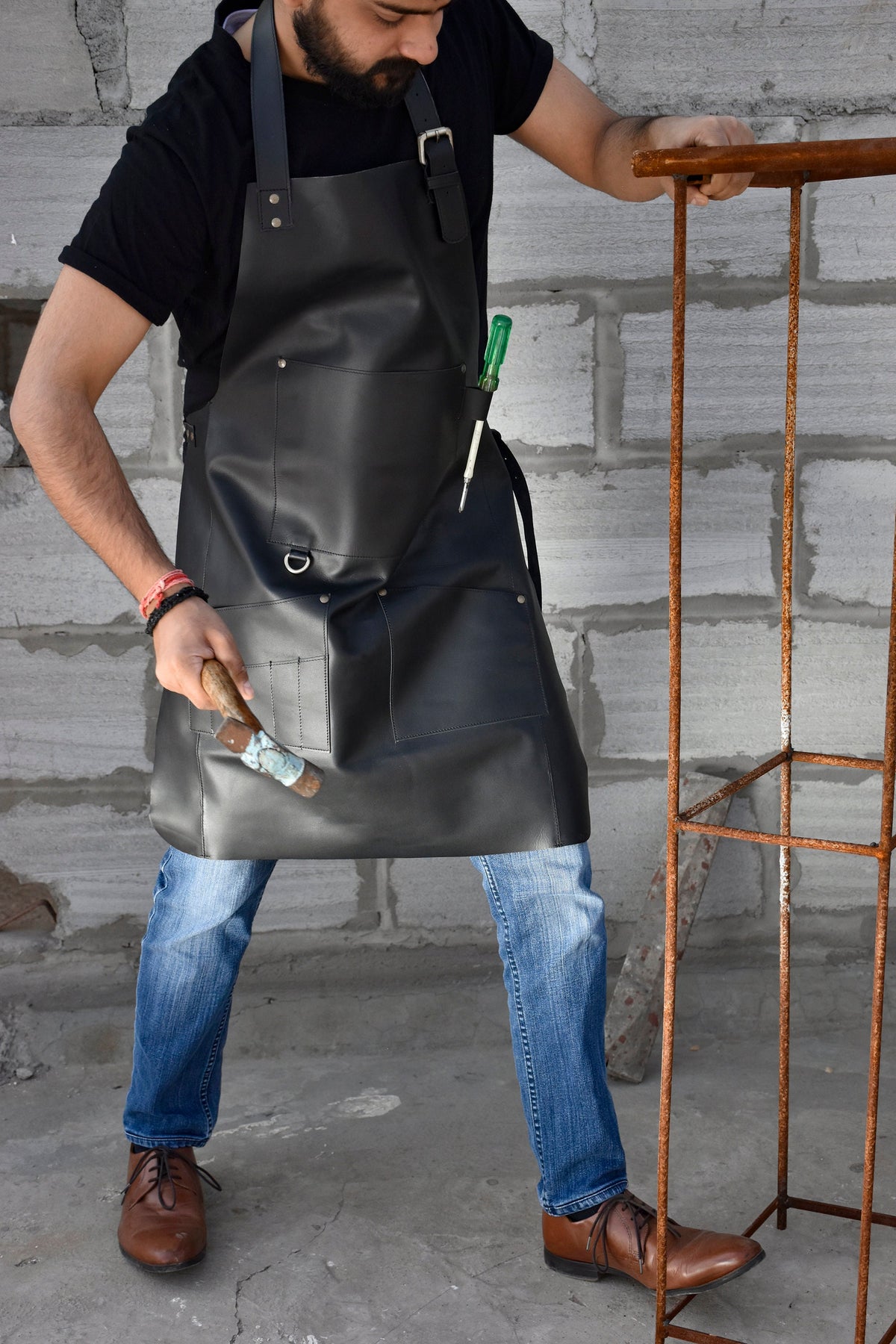 May include: A black leather apron with multiple pockets and a tool holder. The apron is worn by a person with blue jeans and brown shoes.
