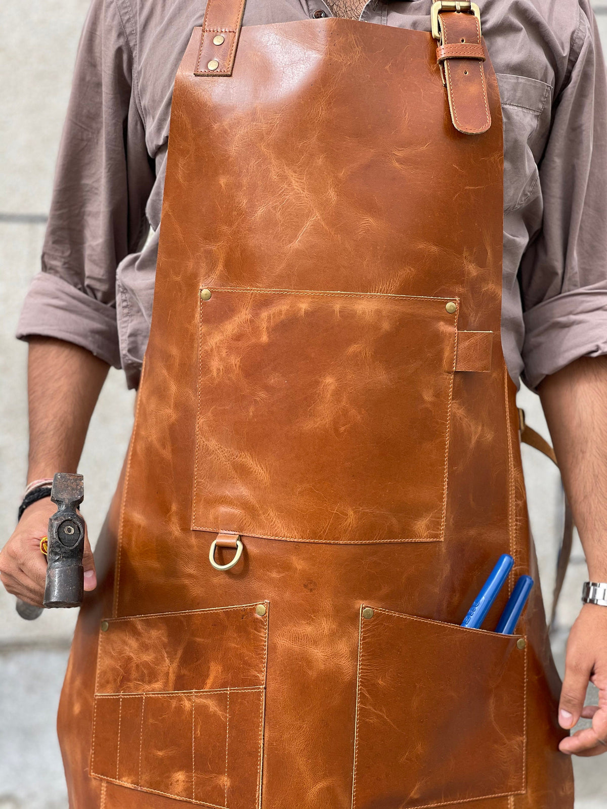 May include: A brown leather apron with multiple pockets, including a large front pocket and a smaller pocket with a metal ring. The apron has a brown leather strap with a metal buckle.