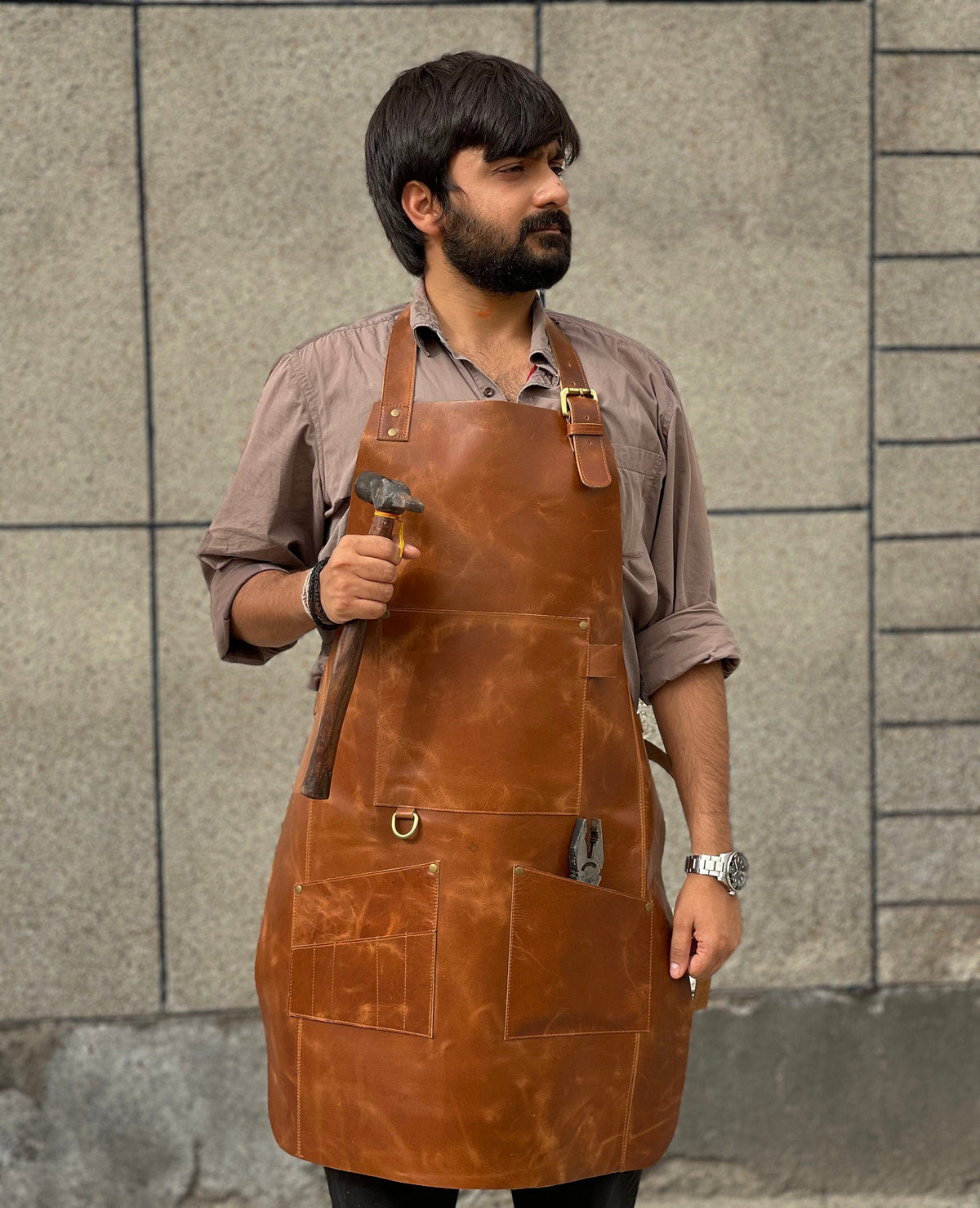 May include: A brown leather apron with multiple pockets and a hammer hanging from the side. The apron has a brass buckle and D-ring.