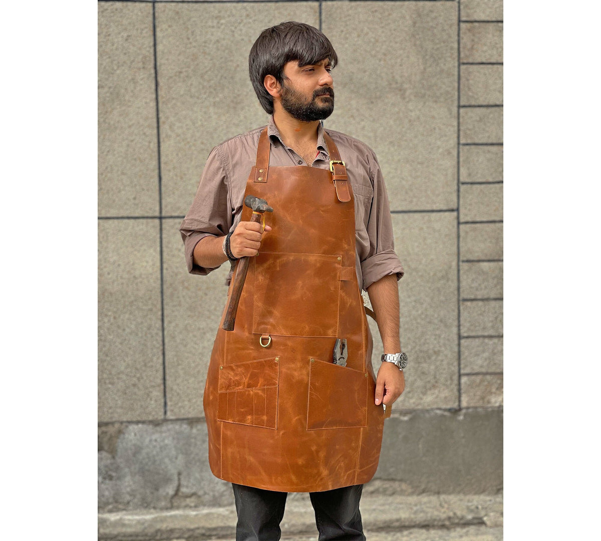 May include: A brown leather apron with multiple pockets and a hammer in the front pocket. The apron has a brass buckle and adjustable straps.