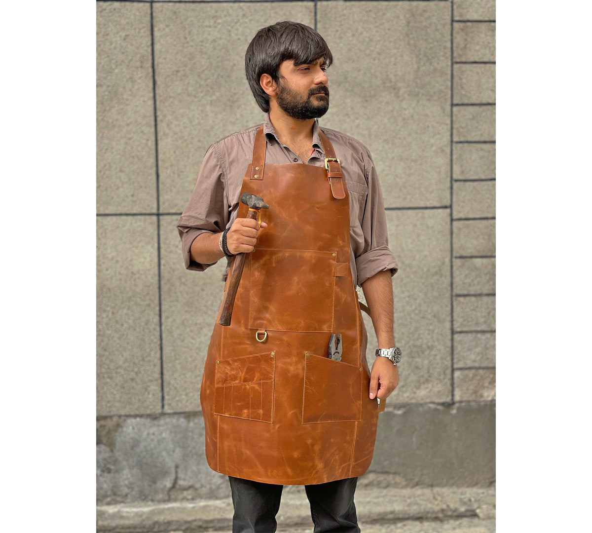 May include: A brown leather apron with multiple pockets and a hammer in the front pocket. The apron has a brass buckle and adjustable straps.