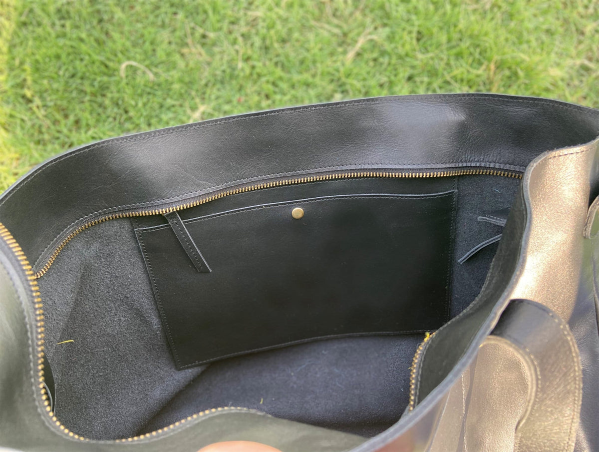 May include: Black leather tote bag with a gold zipper and a small black pocket inside. The bag is open, revealing the interior lining and a small gold button closure. The bag is sitting on green grass.