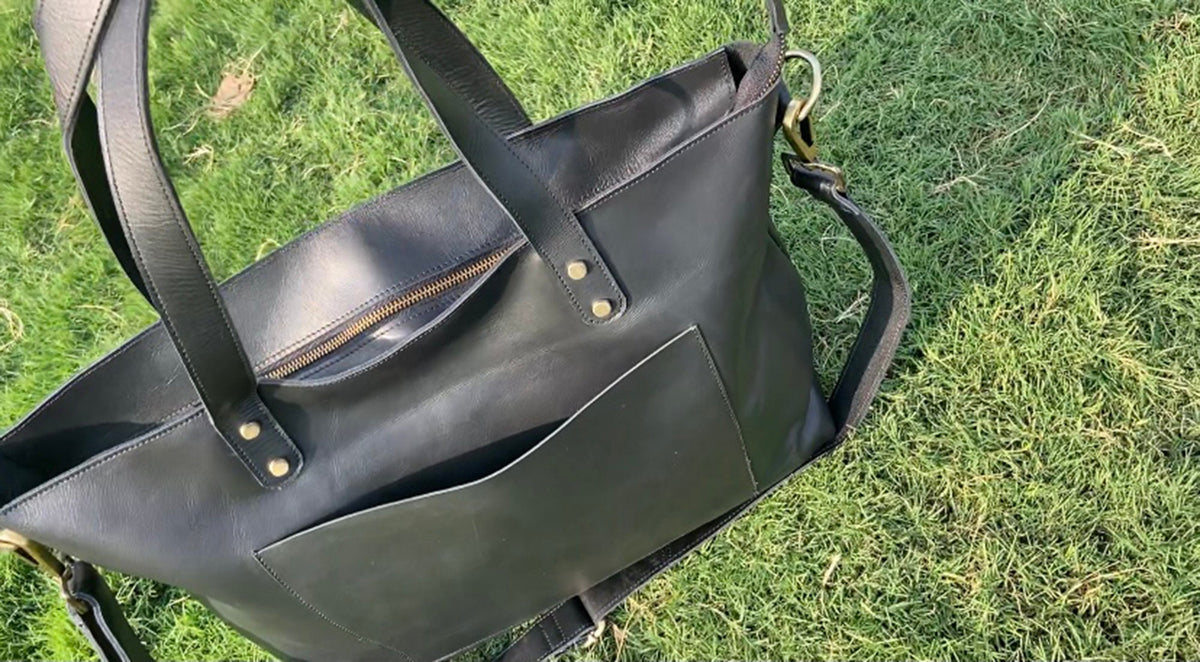 May include: Black leather tote bag with a zippered top and an exterior pocket. The bag has two top handles and a detachable shoulder strap. Gold-colored hardware accents the bag's design. The bag is sitting on a bed of green grass.