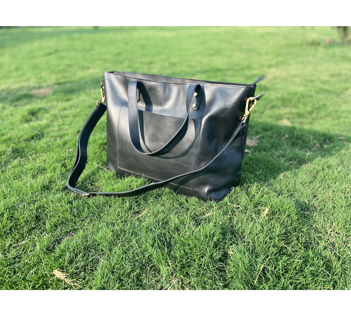 May include: A black leather tote bag with a shoulder strap and top handles, set against a backdrop of green grass. The bag features a front pocket and gold-colored hardware. The bag is approximately 15 inches wide and 12 inches tall.