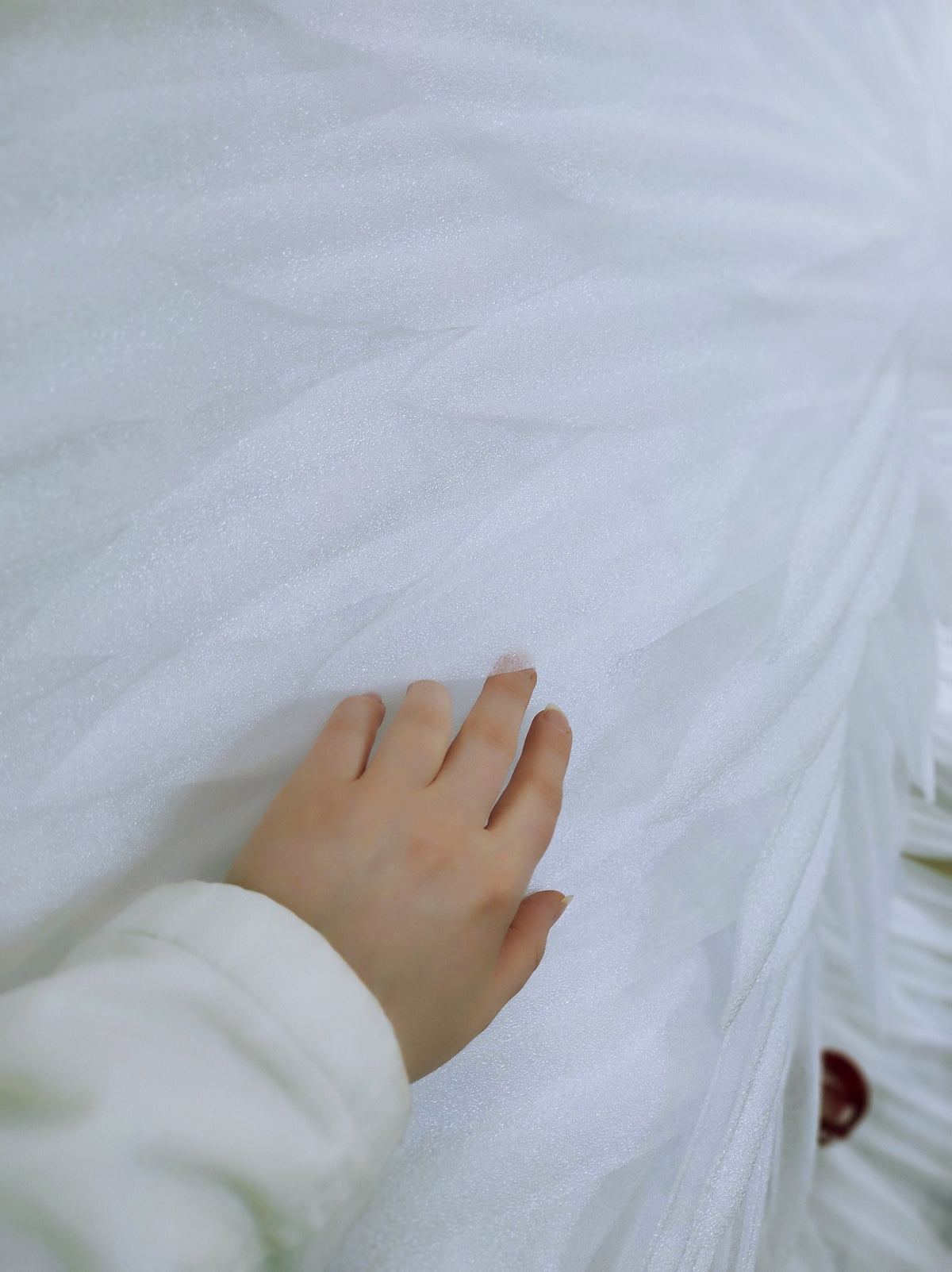 May include: A hand resting on a white tulle fabric. The fabric is soft and delicate, with a slight sheen.