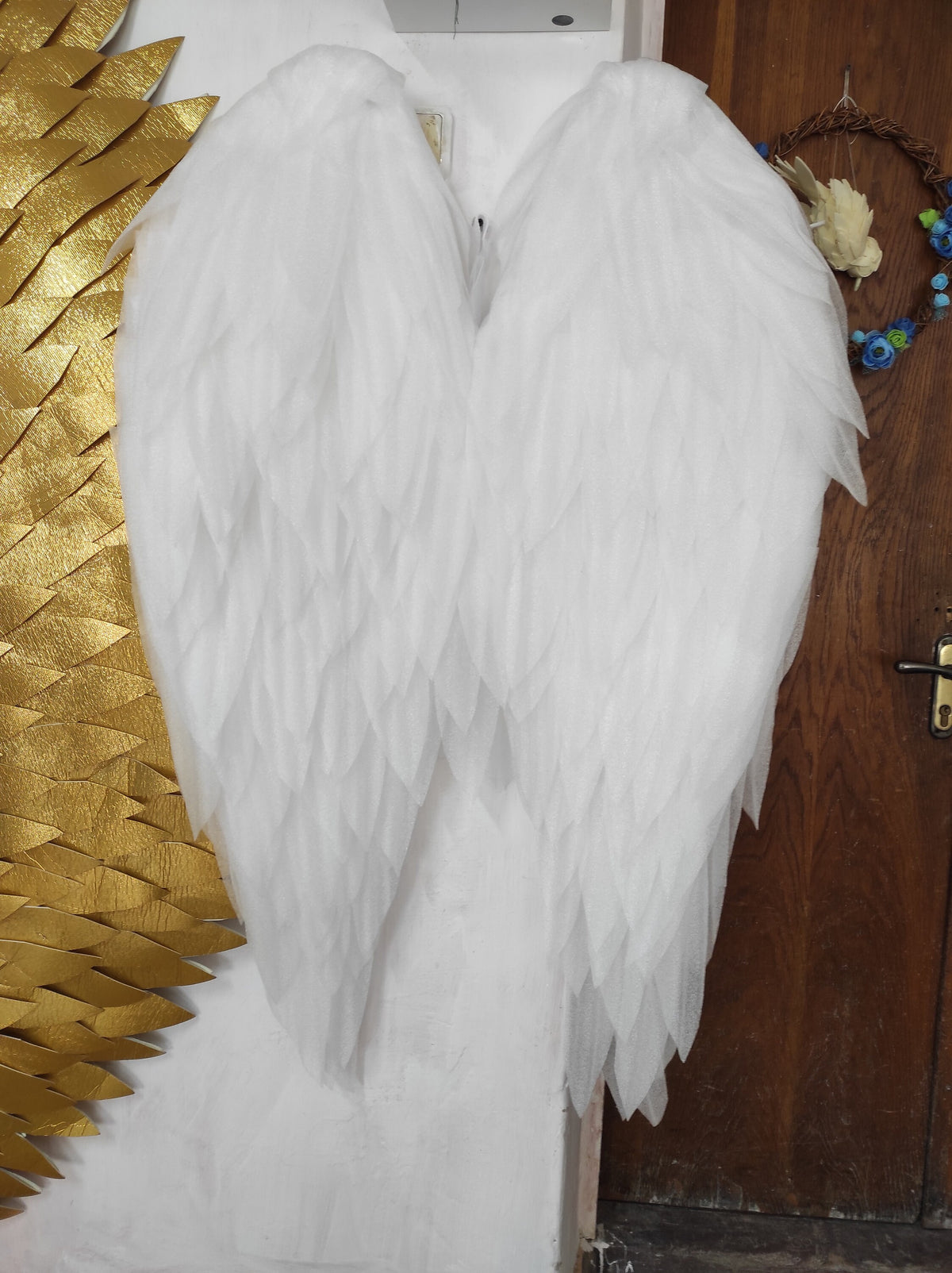 May include: White angel wings made of layered fabric, resembling feathers. The wings are attached to a white background.