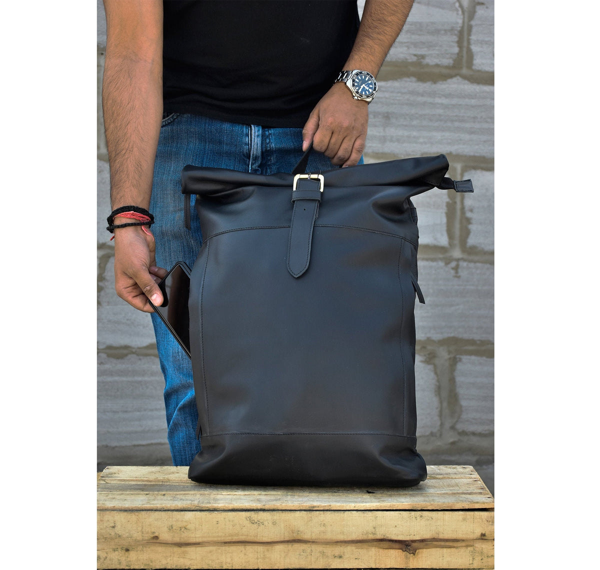 May include: A black leather roll-top backpack with a buckle closure and a strap. The backpack is sitting on a wooden surface.