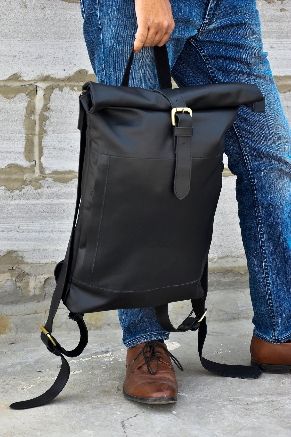 May include: A black leather roll-top backpack with a gold buckle and adjustable straps. The backpack is being held by a person wearing blue jeans.