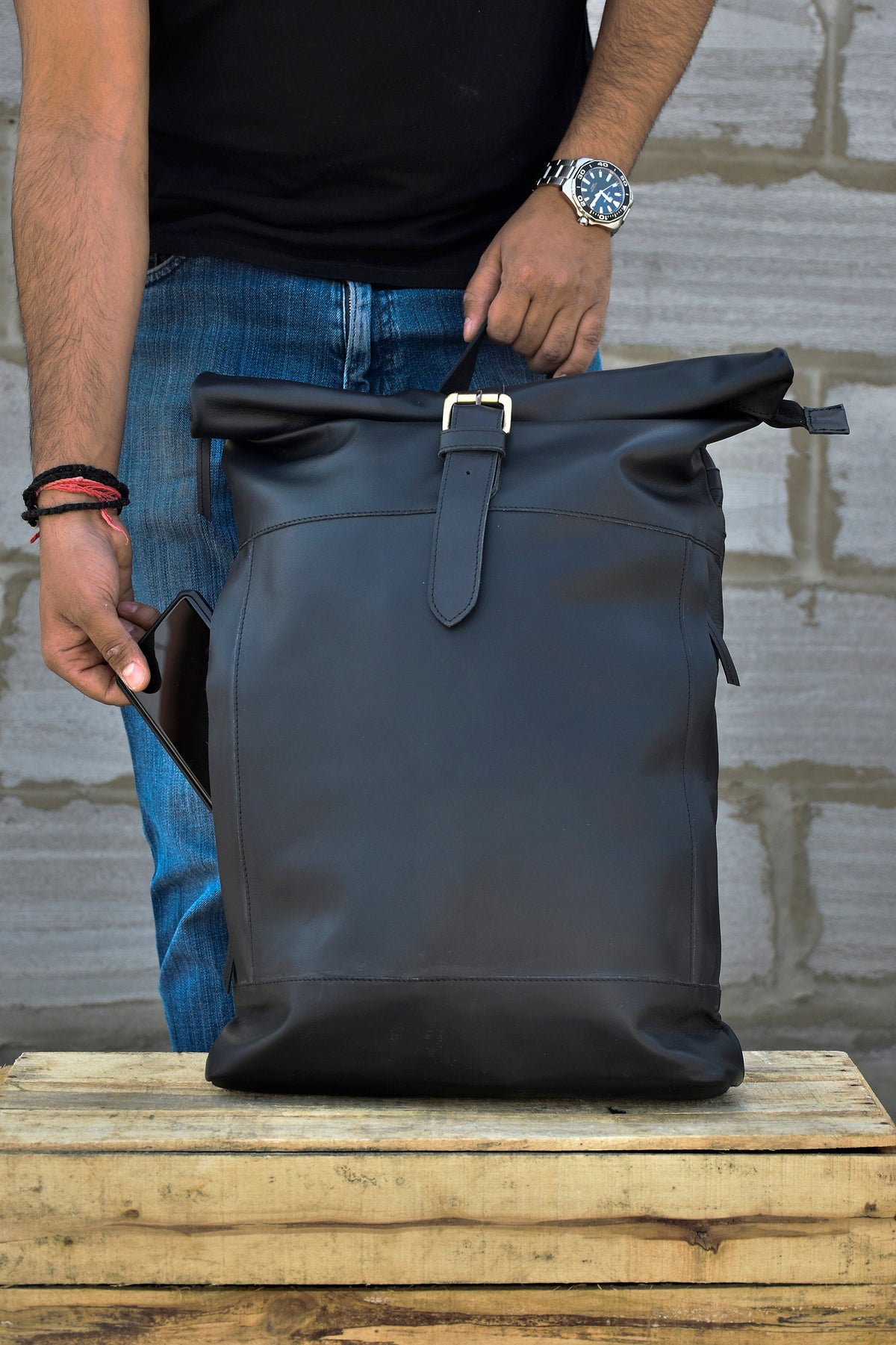 May include: A black leather roll-top backpack with a buckle closure and a side pocket. The backpack is sitting on a wooden crate.