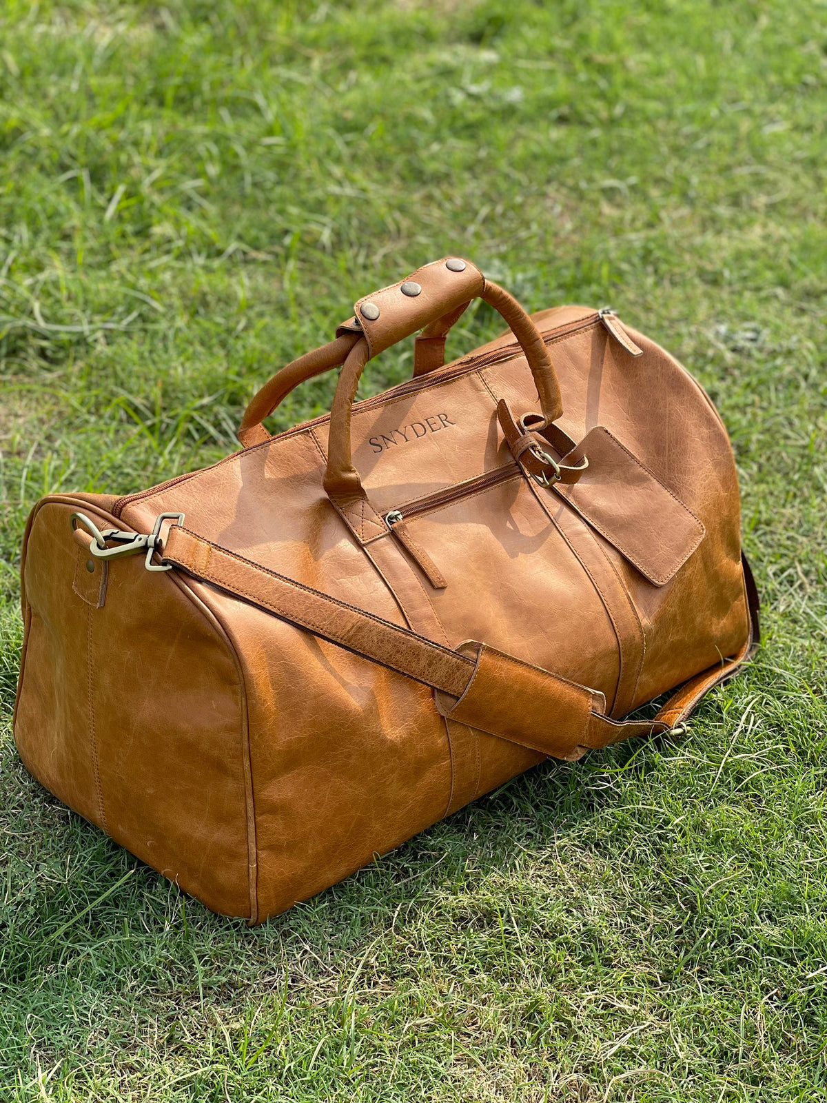 May include: A tan leather duffel bag featuring the name SNYDER embossed on the front, two top handles with a snap-button wrap, a detachable shoulder strap with a padded section, a front zippered pocket, and an attached matching luggage tag.