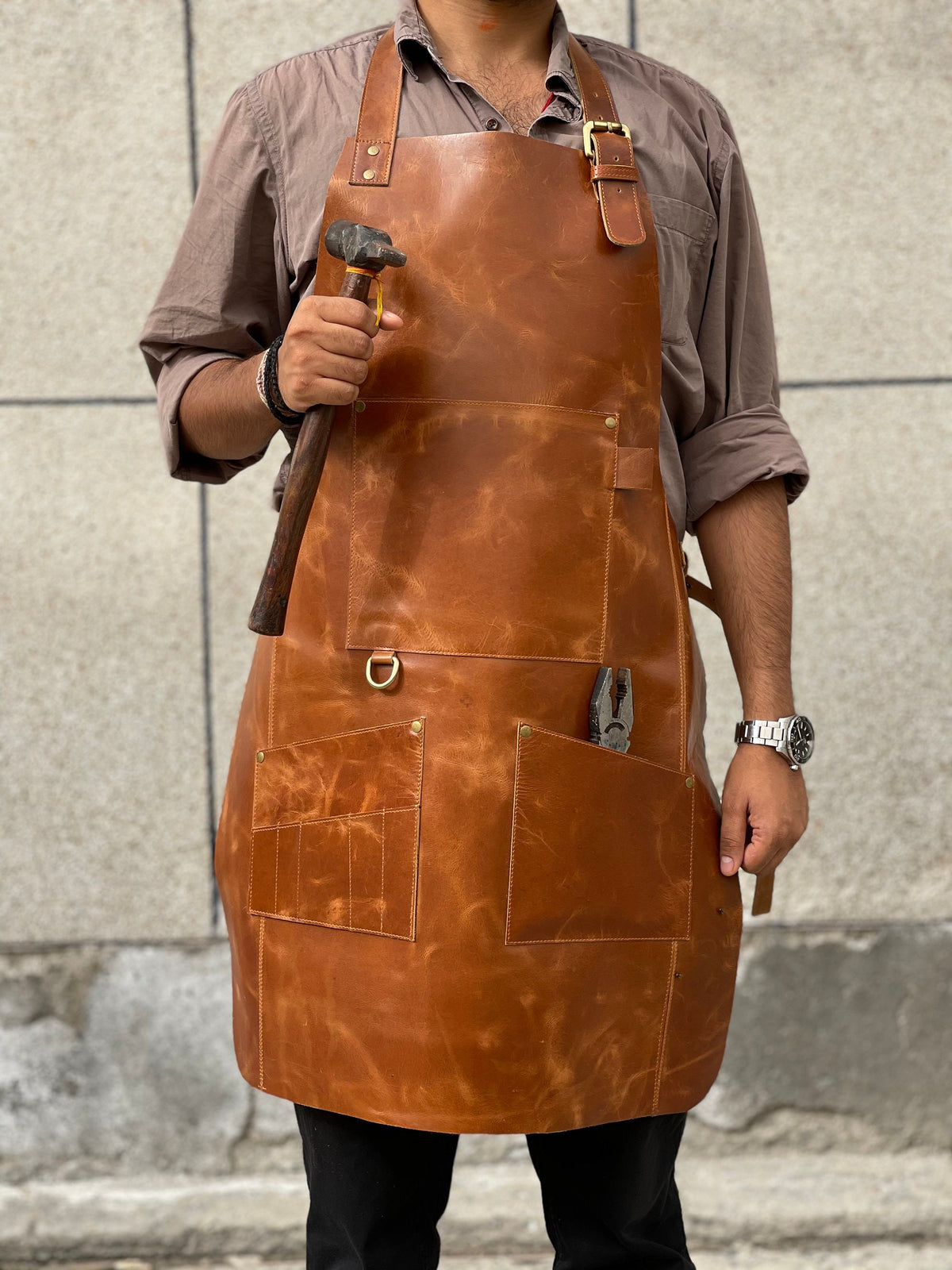 May include: A brown leather apron with multiple pockets and a hammer. The apron has a strap that goes around the neck and another strap that goes around the waist.