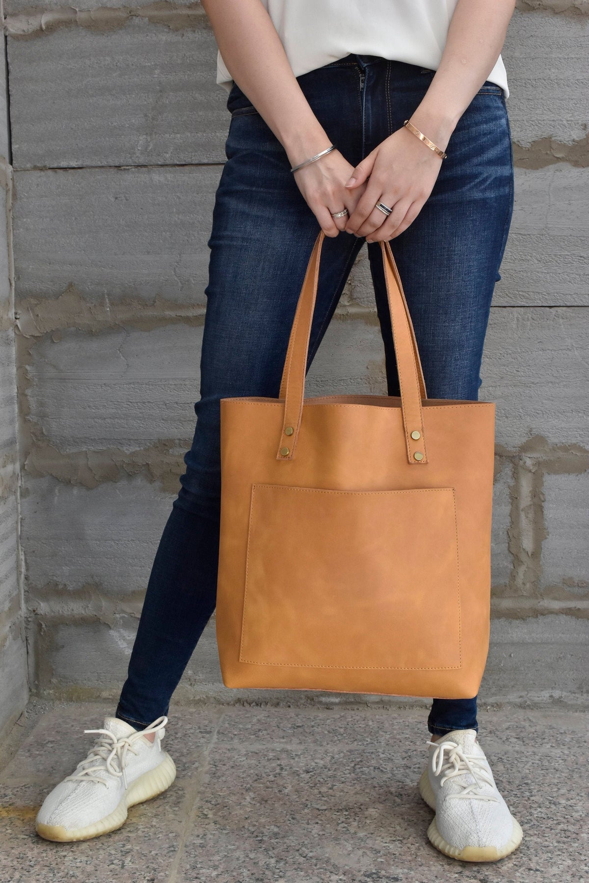 May include: A tan leather tote bag with a front pocket and gold hardware. The bag is being held by a person wearing blue jeans and white sneakers.