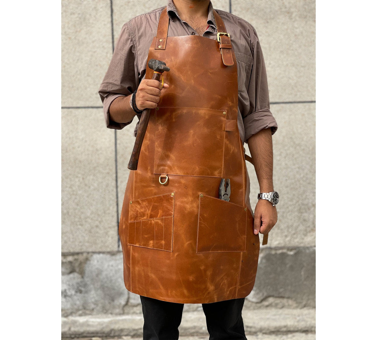 May include: A brown leather apron with multiple pockets and a hammer and pliers in the pockets. The apron has a brass buckle and a brass ring.