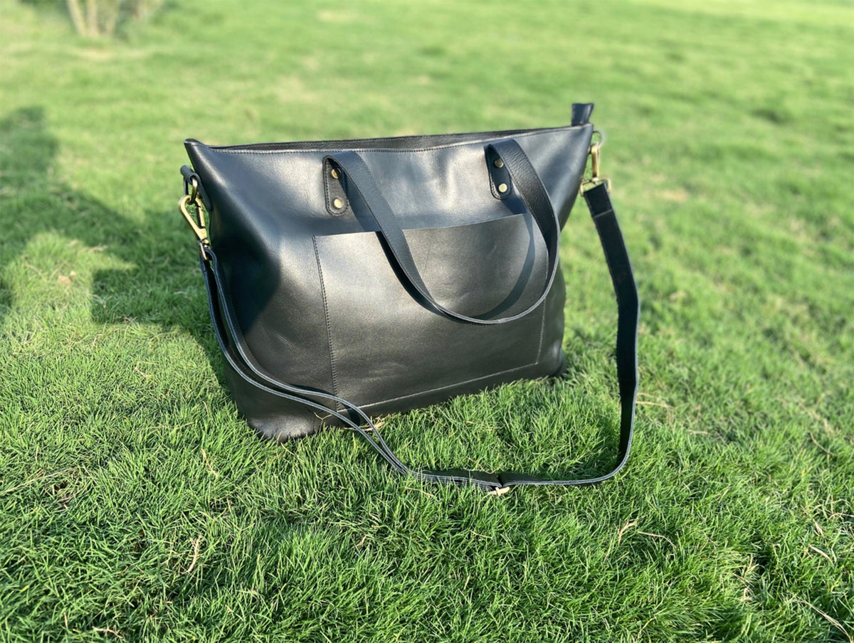 May include: A black leather tote bag with a shoulder strap and a front pocket. The bag has gold-colored hardware and is sitting on a bed of green grass. The bag has two handles and a long, adjustable strap.
