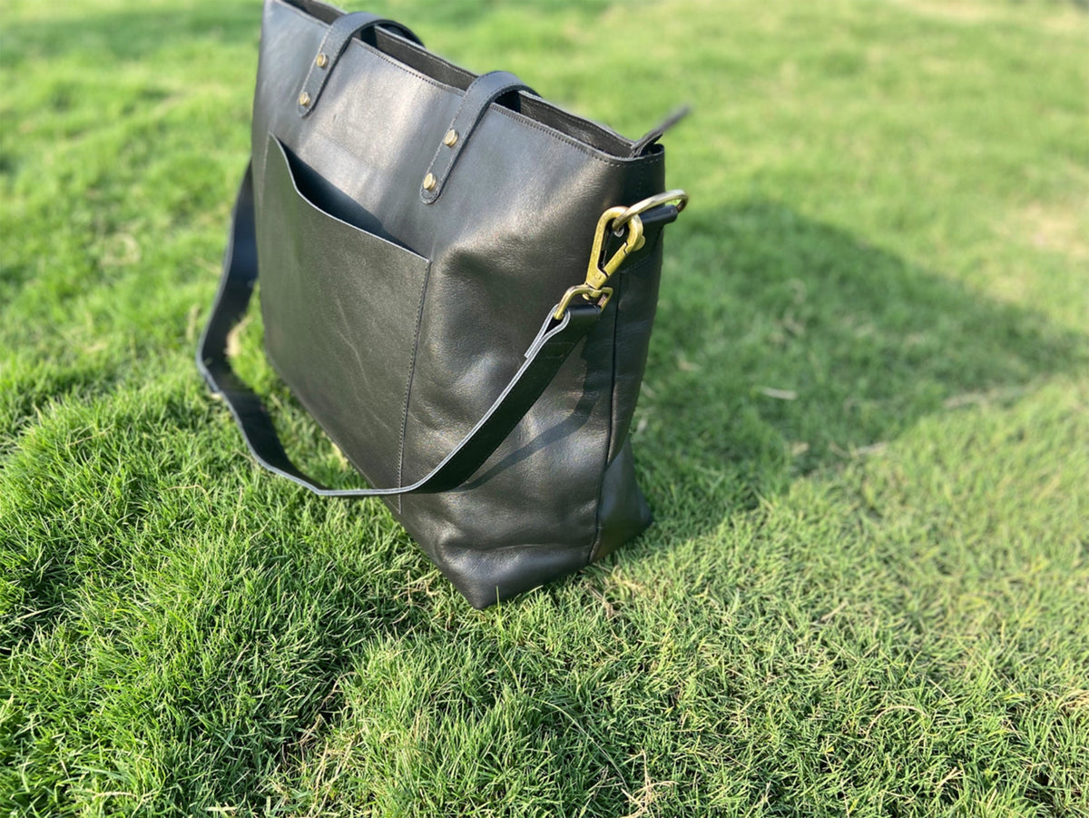 May include: Black leather tote bag with a front pocket and a detachable shoulder strap. The bag has brass-colored hardware and is sitting on green grass. The tote bag has two top handles and is designed for carrying items.