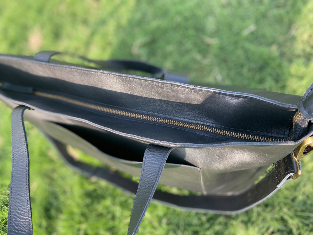 May include: A black leather tote bag with a zipper closure and a shoulder strap. The bag has a large interior pocket and a smaller exterior pocket. The hardware is gold-colored. The bag is sitting on a green grassy surface.