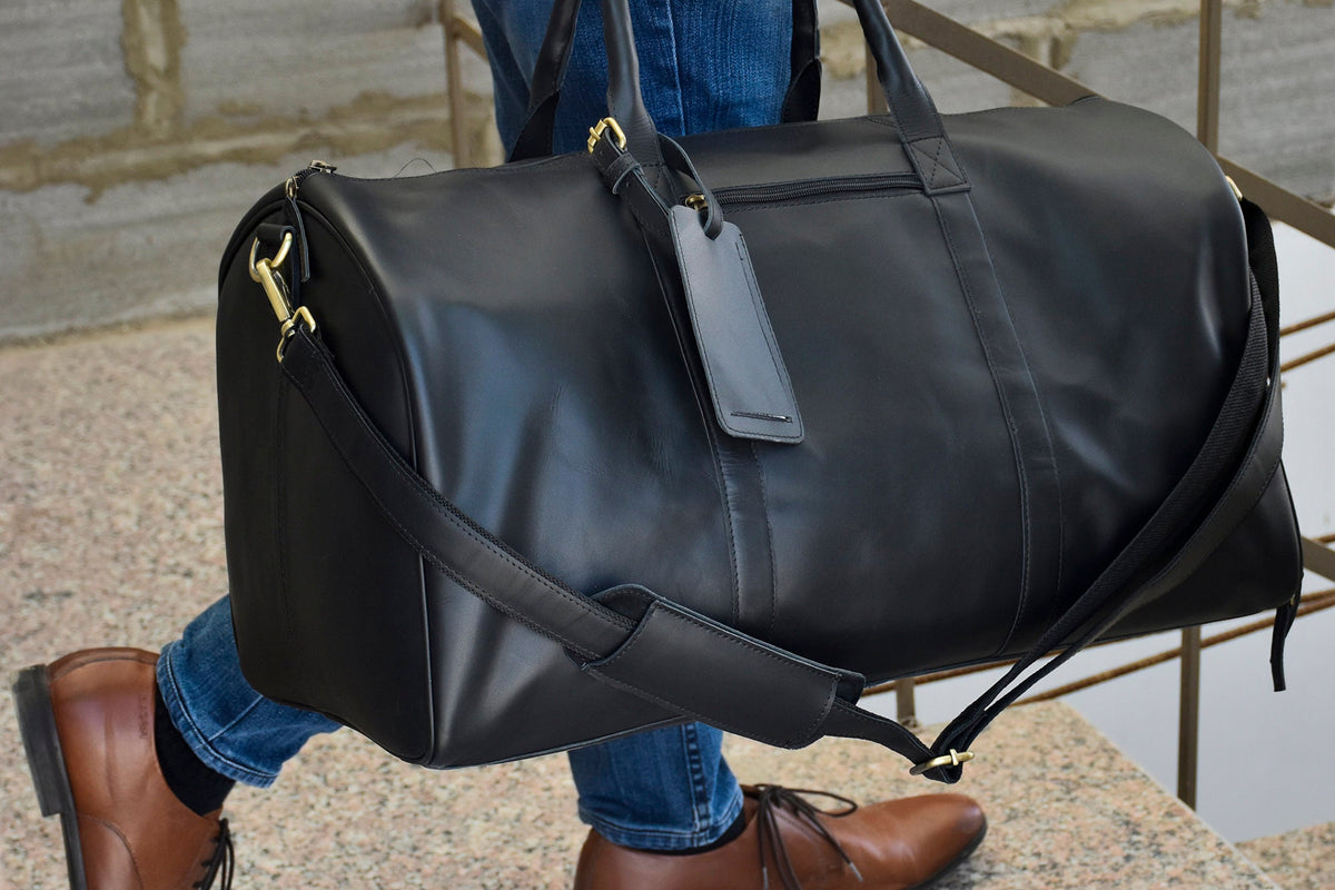 May include: A black leather duffel bag with a gold zipper and a gold luggage tag. The bag has a long strap that can be worn over the shoulder.