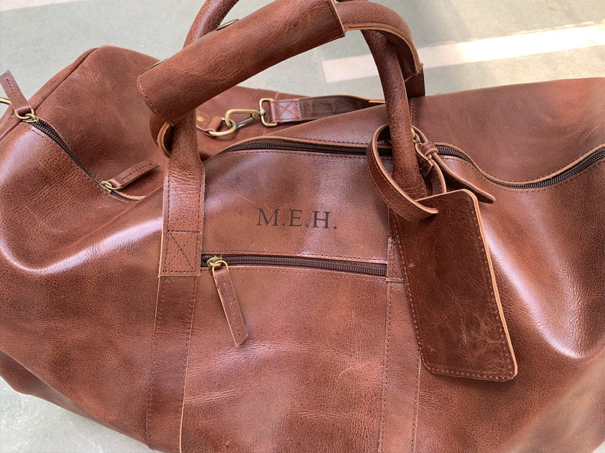 May include: A brown leather duffel bag with a zipper closure and a leather handle. The bag has a luggage tag and the initials "M.E.H." are embossed on the front.