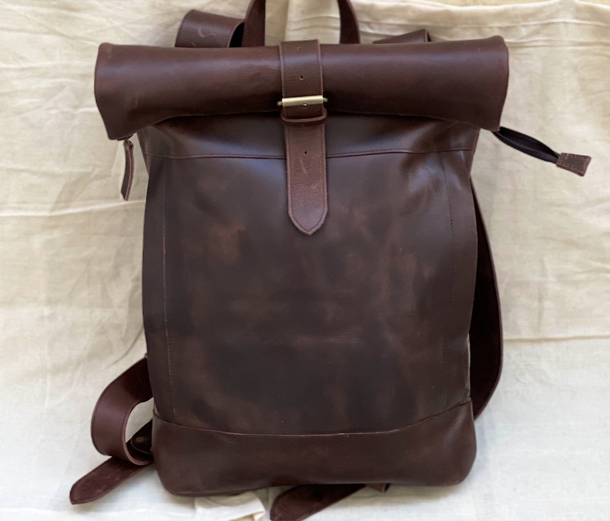 May include: A brown leather backpack with a roll-top closure and a buckle. The backpack has a distressed look and a single strap.