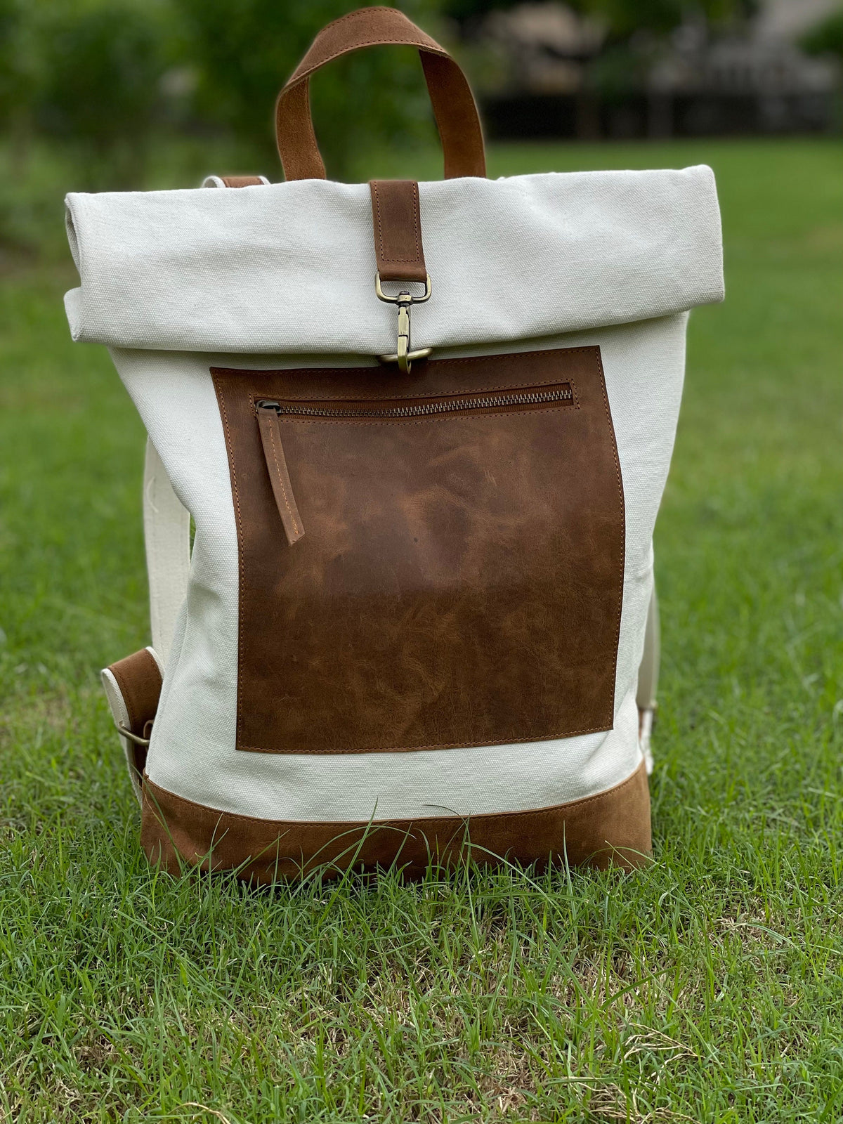 May include: A white canvas backpack with a brown leather flap and a large brown leather pocket on the front. The backpack has a roll-top closure and a leather strap.