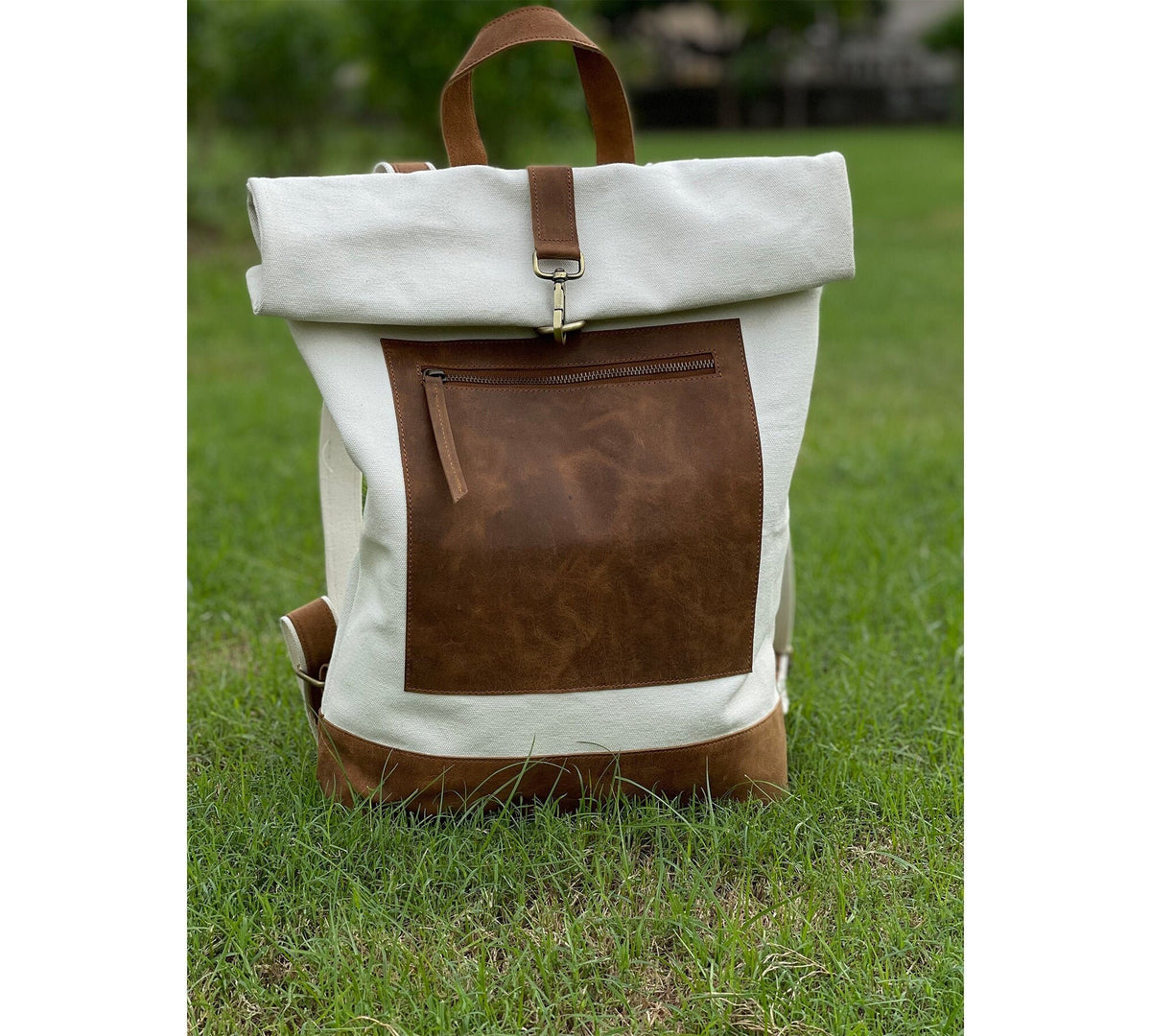 May include: A white canvas backpack with a brown leather flap and a zippered pocket. The backpack has a roll-top closure and adjustable straps.