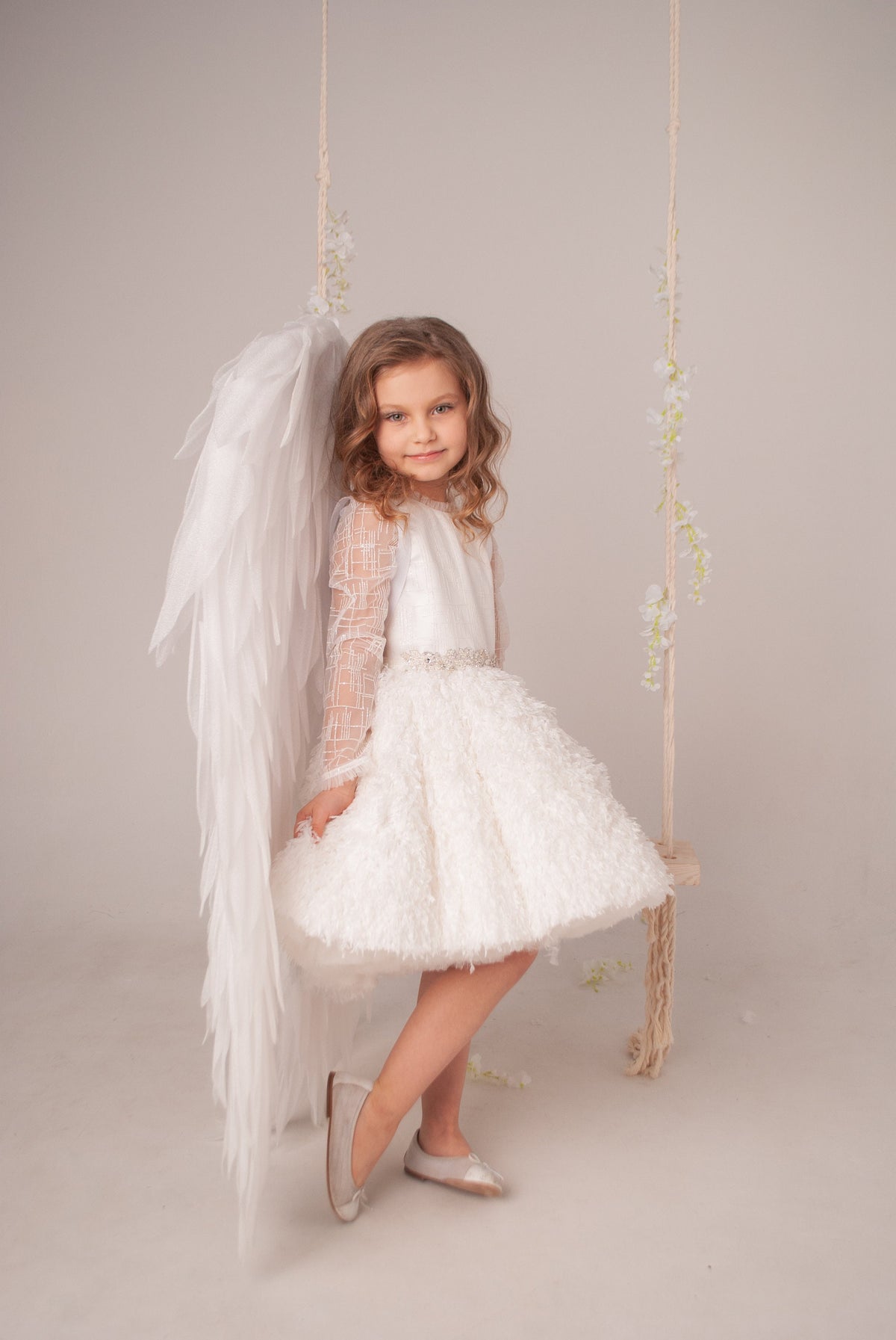 May include: A white dress with long sheer sleeves featuring a grid pattern, a textured skirt, and a beaded waistline. Large white feathered angel wings are attached to the back. The outfit is paired with simple light-colored ballet flats.