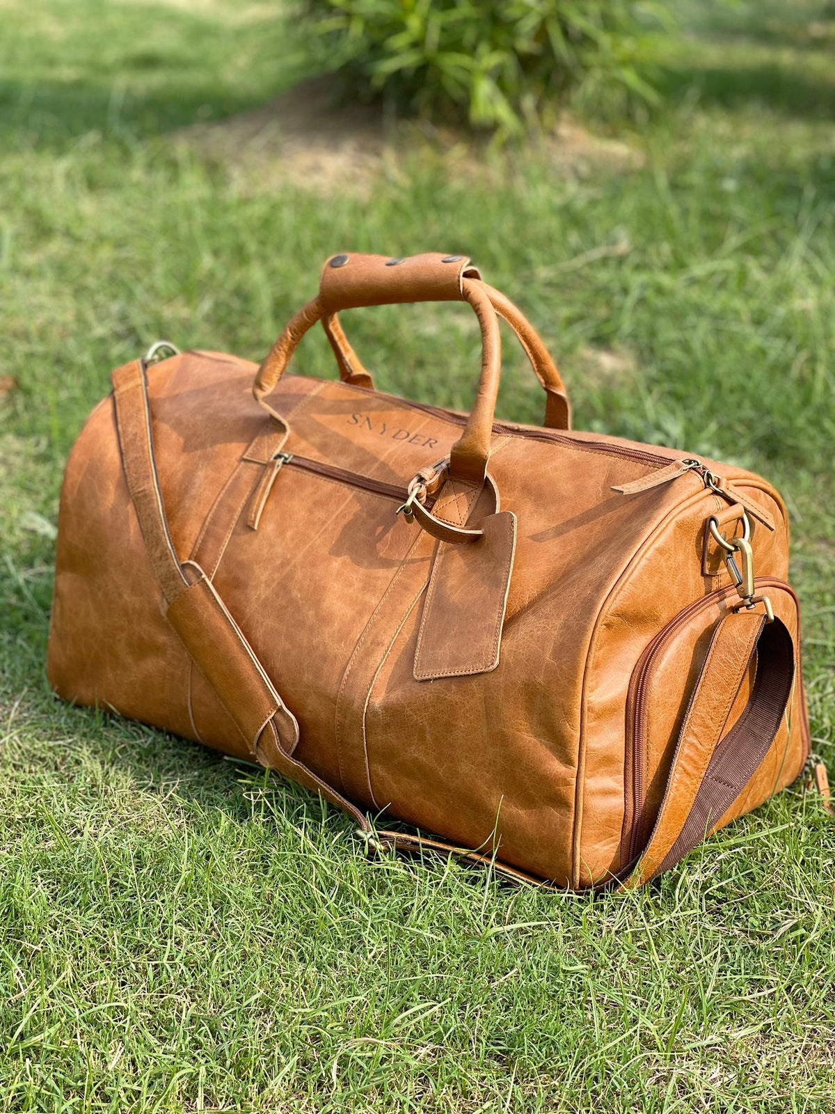 May include: A brown leather duffel bag with a top handle and a shoulder strap. The bag has a zipper closure and a small pocket on the side. The bag is personalized with the name "Snyder" embossed on the front.