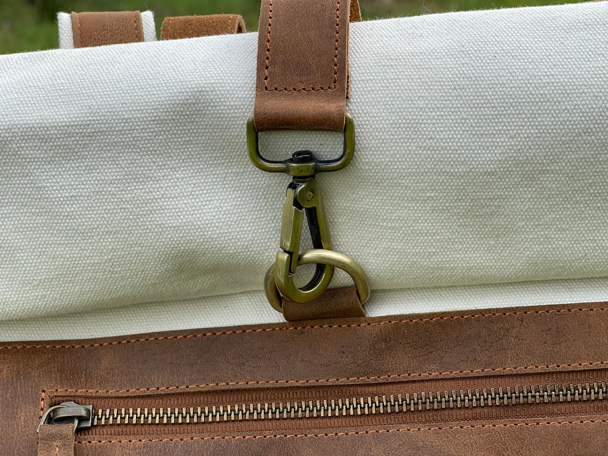 May include: Close-up of a white canvas backpack with a brown leather strap and a brass hook closure. The backpack has a brown leather zippered pocket.