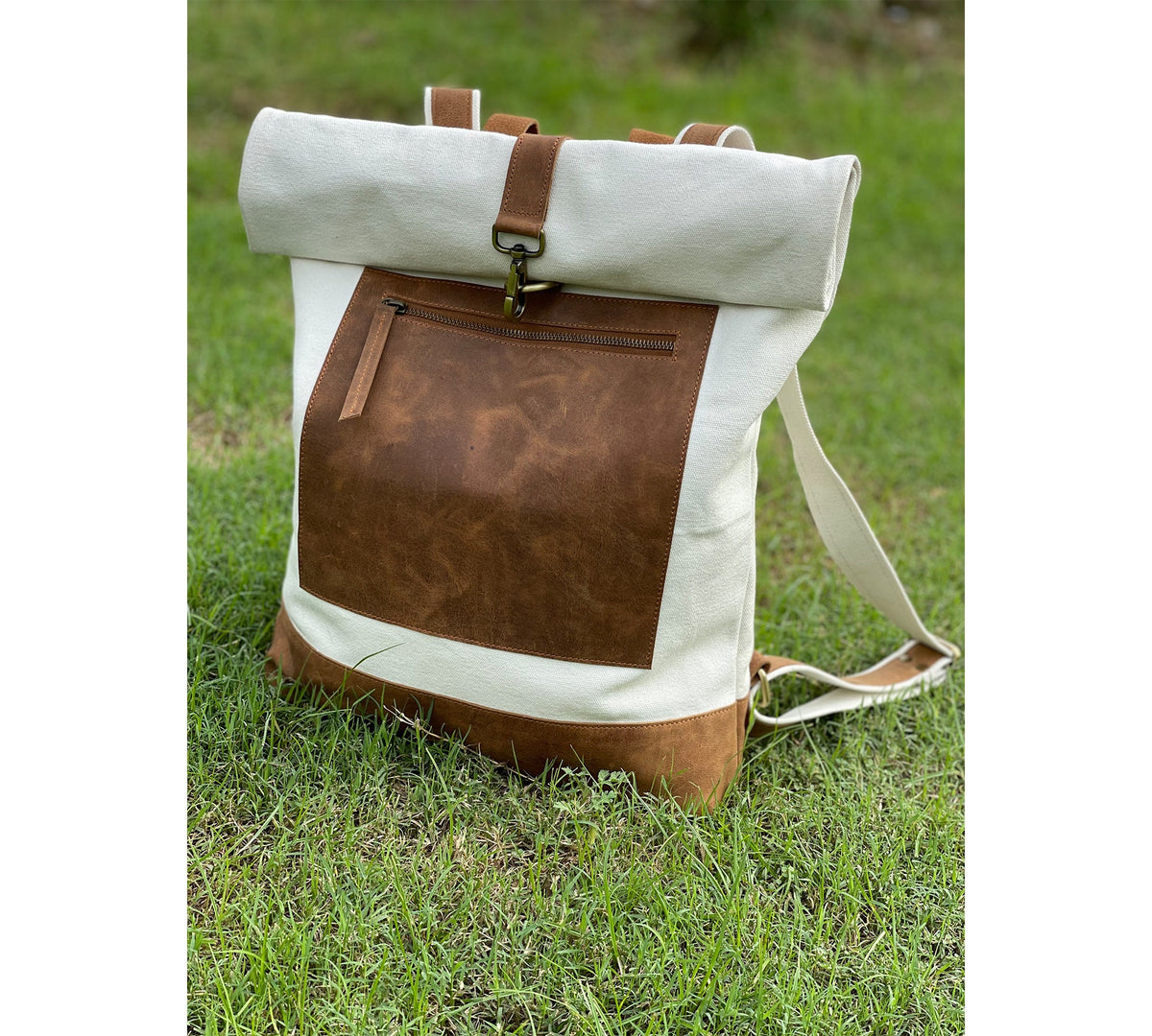 May include: A white canvas backpack with a brown leather flap and a zippered pocket. The backpack has a roll-top closure and adjustable straps.